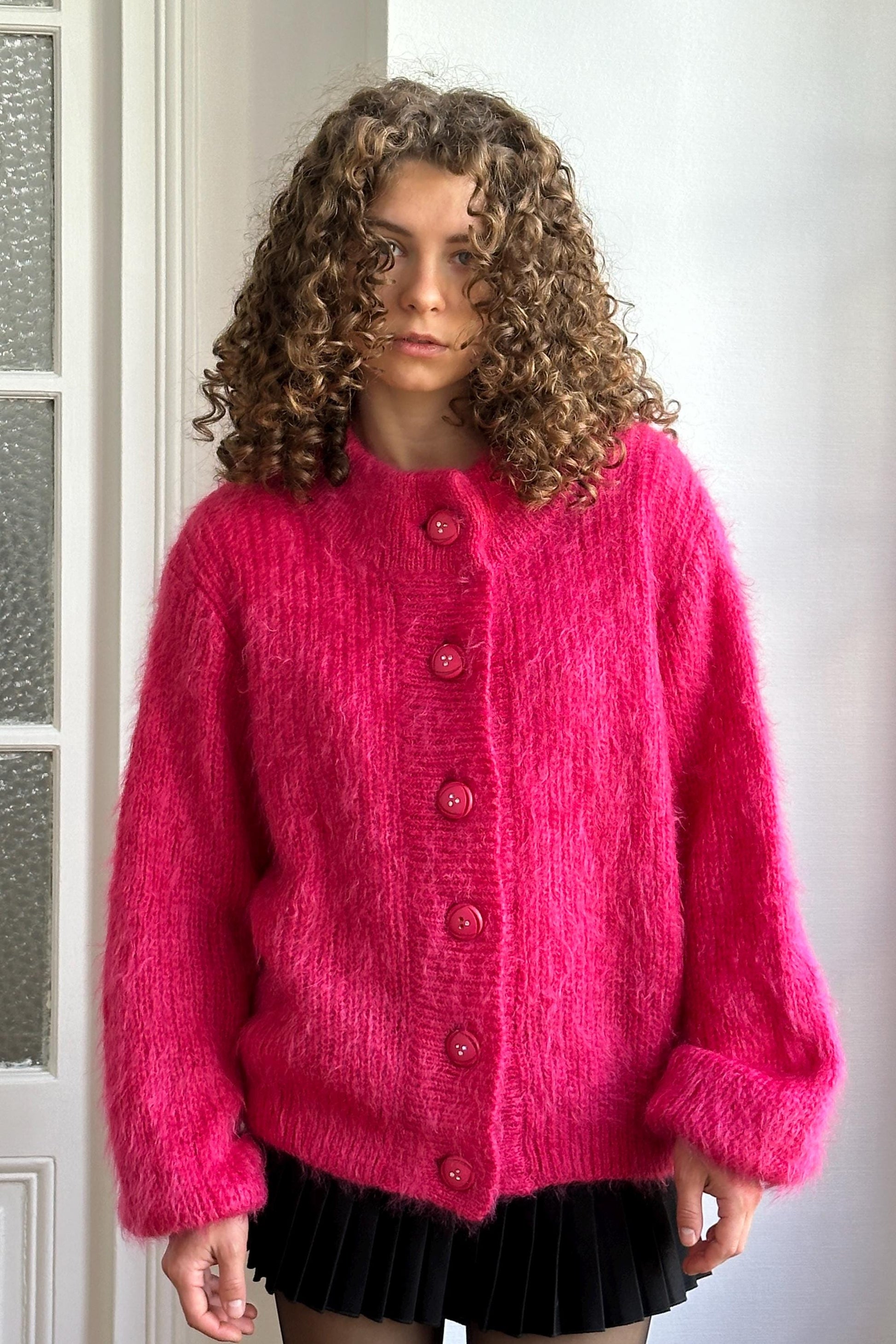 A woman with curly hair wearing a bright pink cardigan and black skirt stands in front of a white door.
