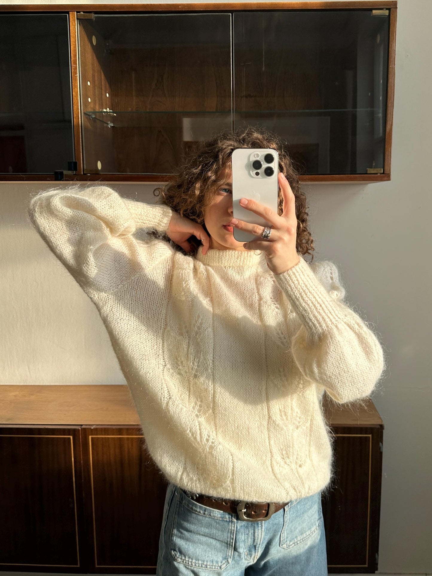 A person is taking a selfie with a white iPhone, wearing a white sweater and jeans, in front of a wooden cabinet.