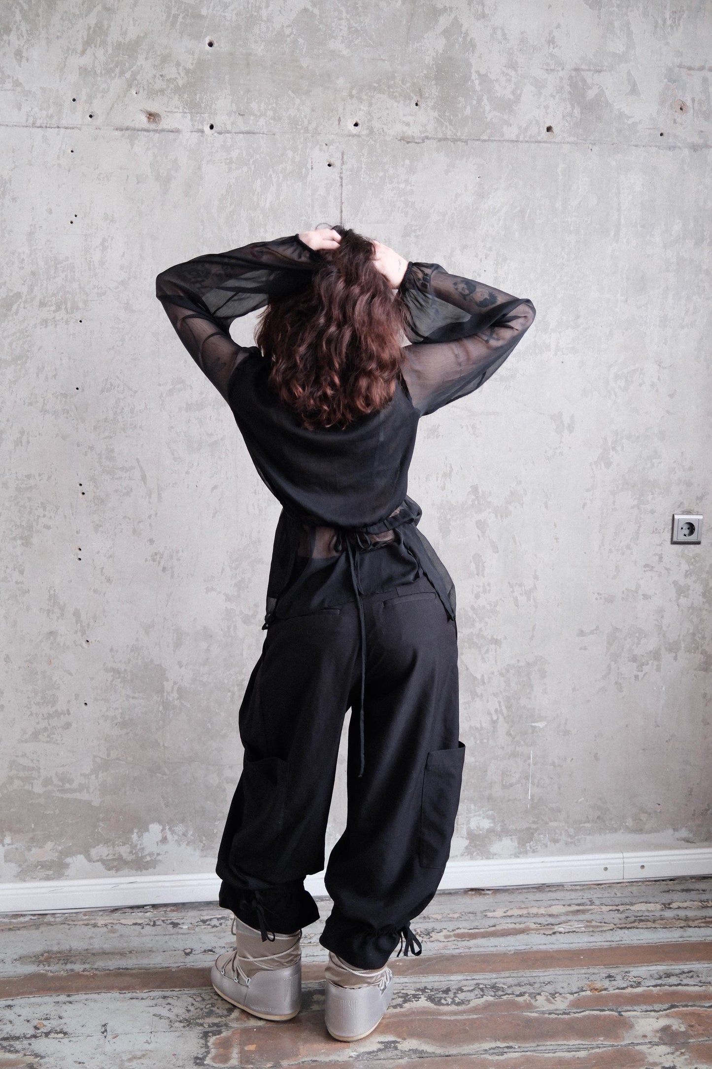 A person in a black outfit stands in front of a concrete wall, with their hands on their head.