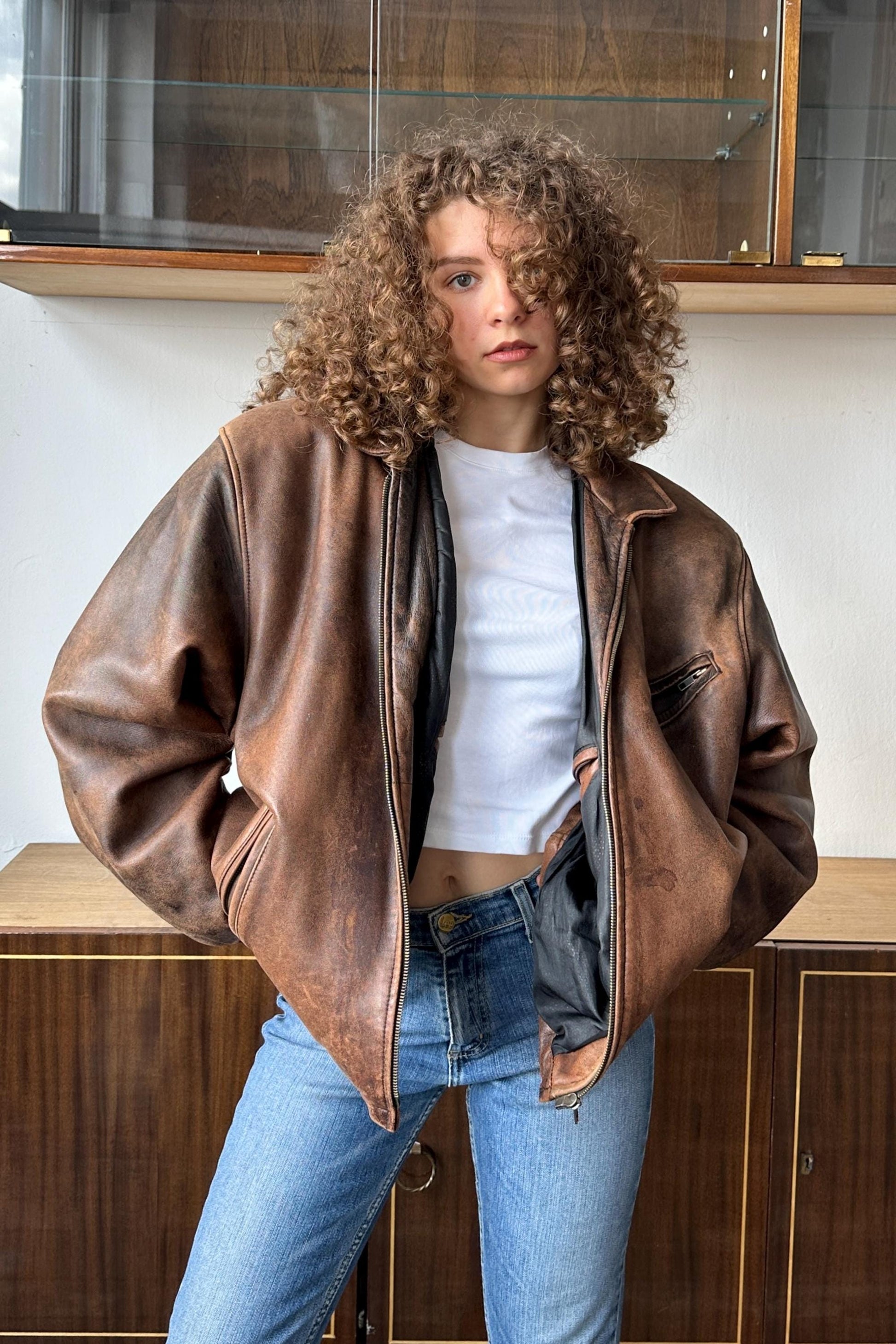 A young woman with curly hair wearing a brown leather jacket, white shirt, and blue jeans stands in front of a wooden cabinet.