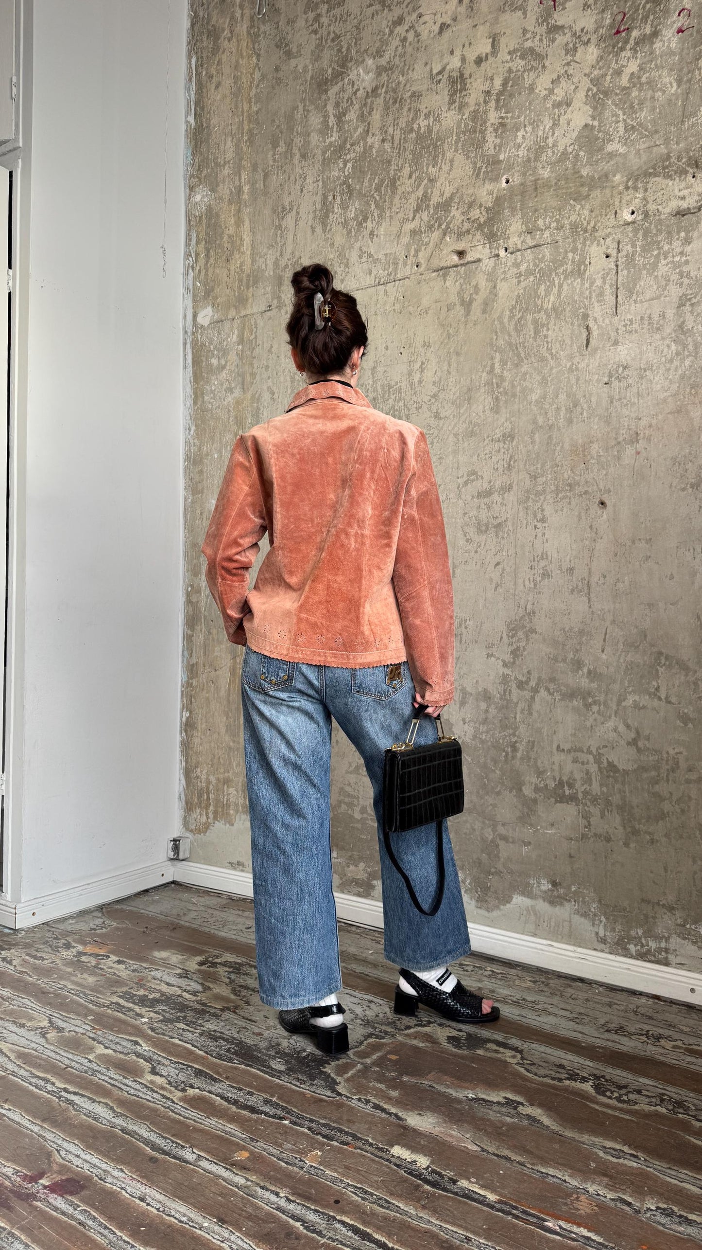 A person wearing a pink jacket and blue jeans stands in front of a concrete wall, holding a black purse.