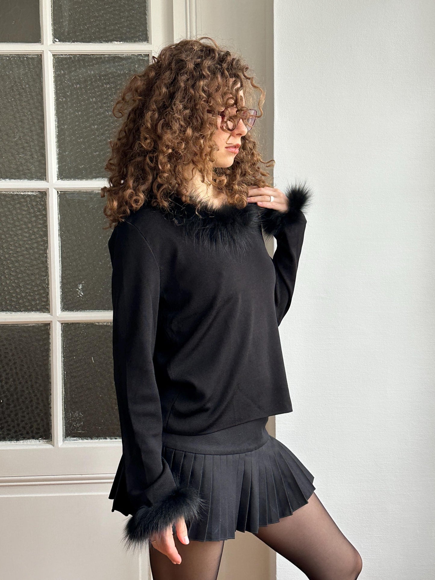 A woman with curly hair wearing a black top and skirt stands in front of a window.
