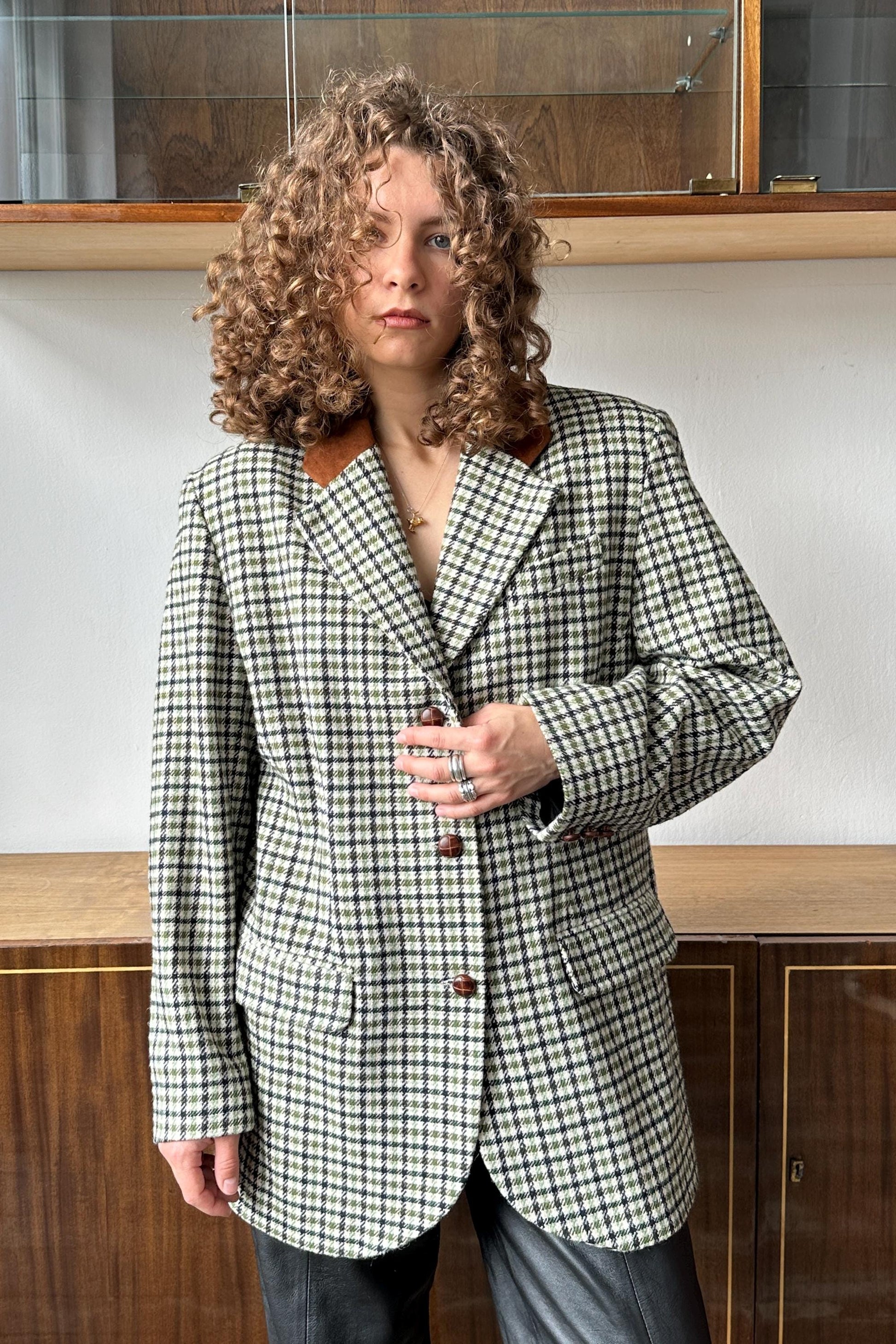 A woman with curly hair wearing a black and white checkered blazer stands in front of a wooden cabinet.
