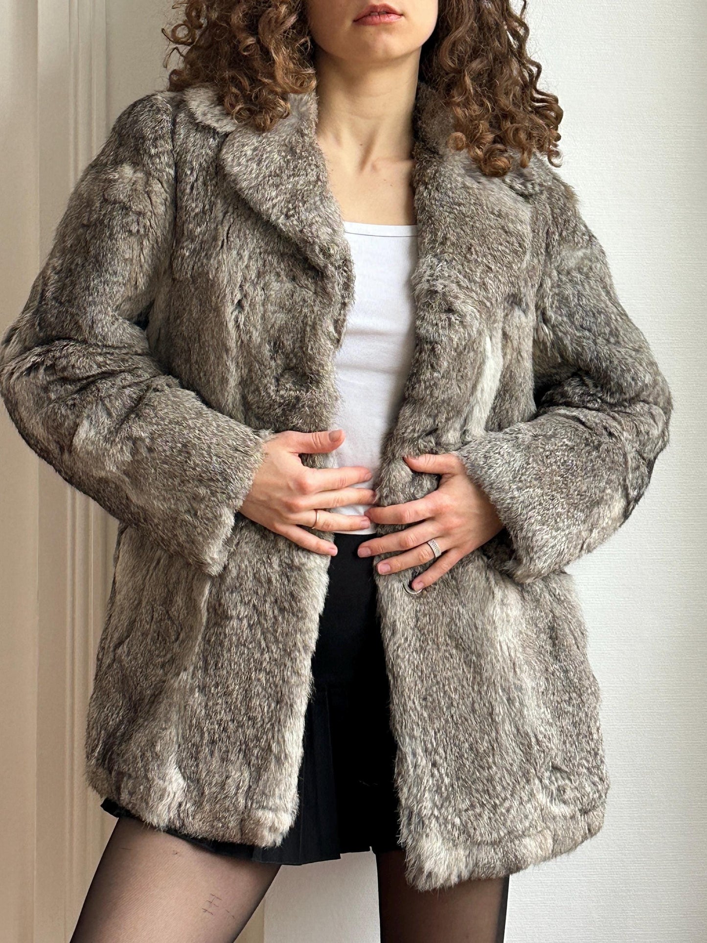 A woman wearing a gray fur coat and black skirt stands in front of a white wall.