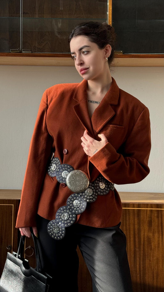 A woman wearing a brown blazer and holding a large belt with a circular design, posing in front of a wooden wall.
