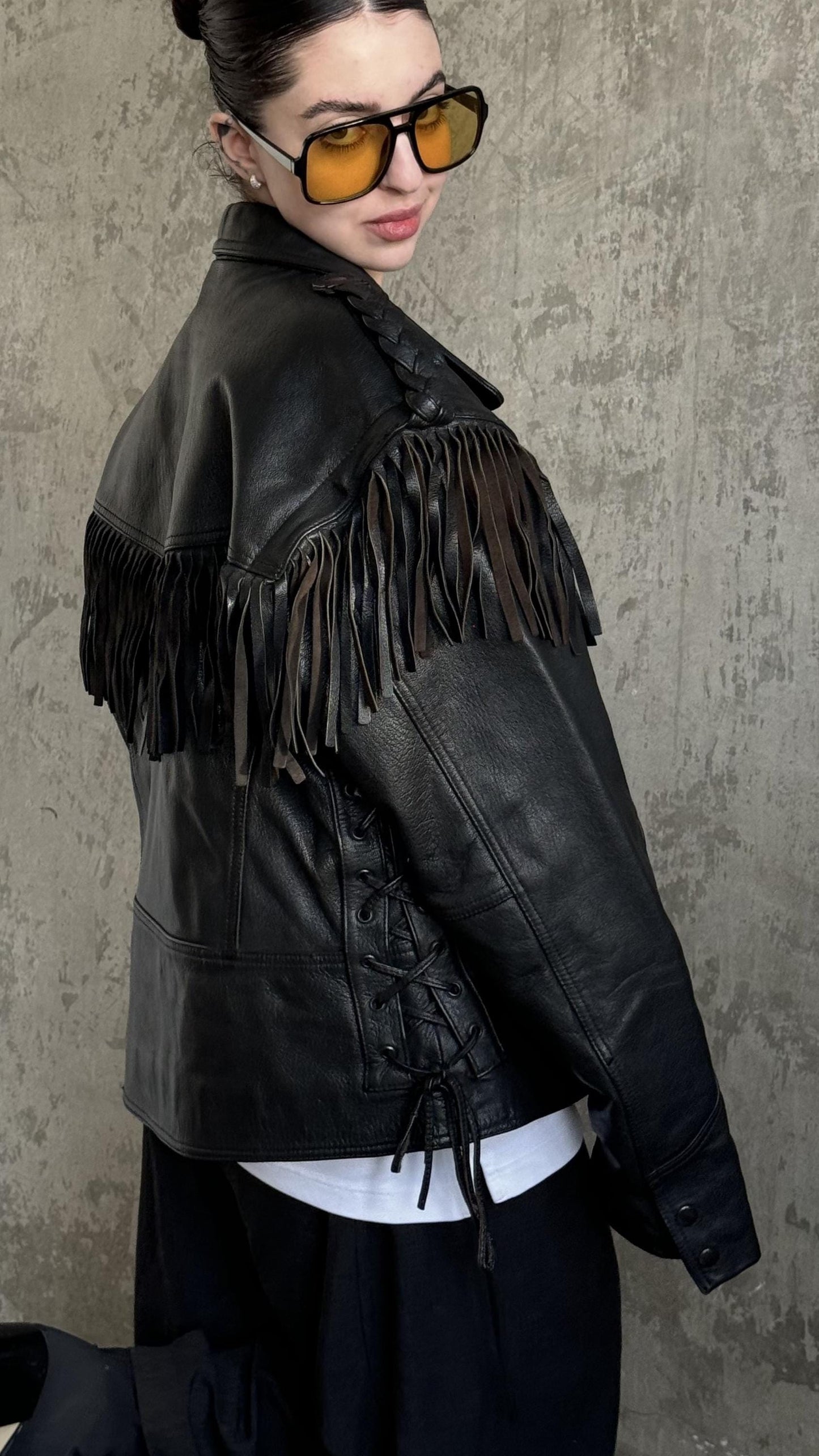 A woman wearing a black leather jacket with fringe, sunglasses, and a white shirt stands against a concrete wall.