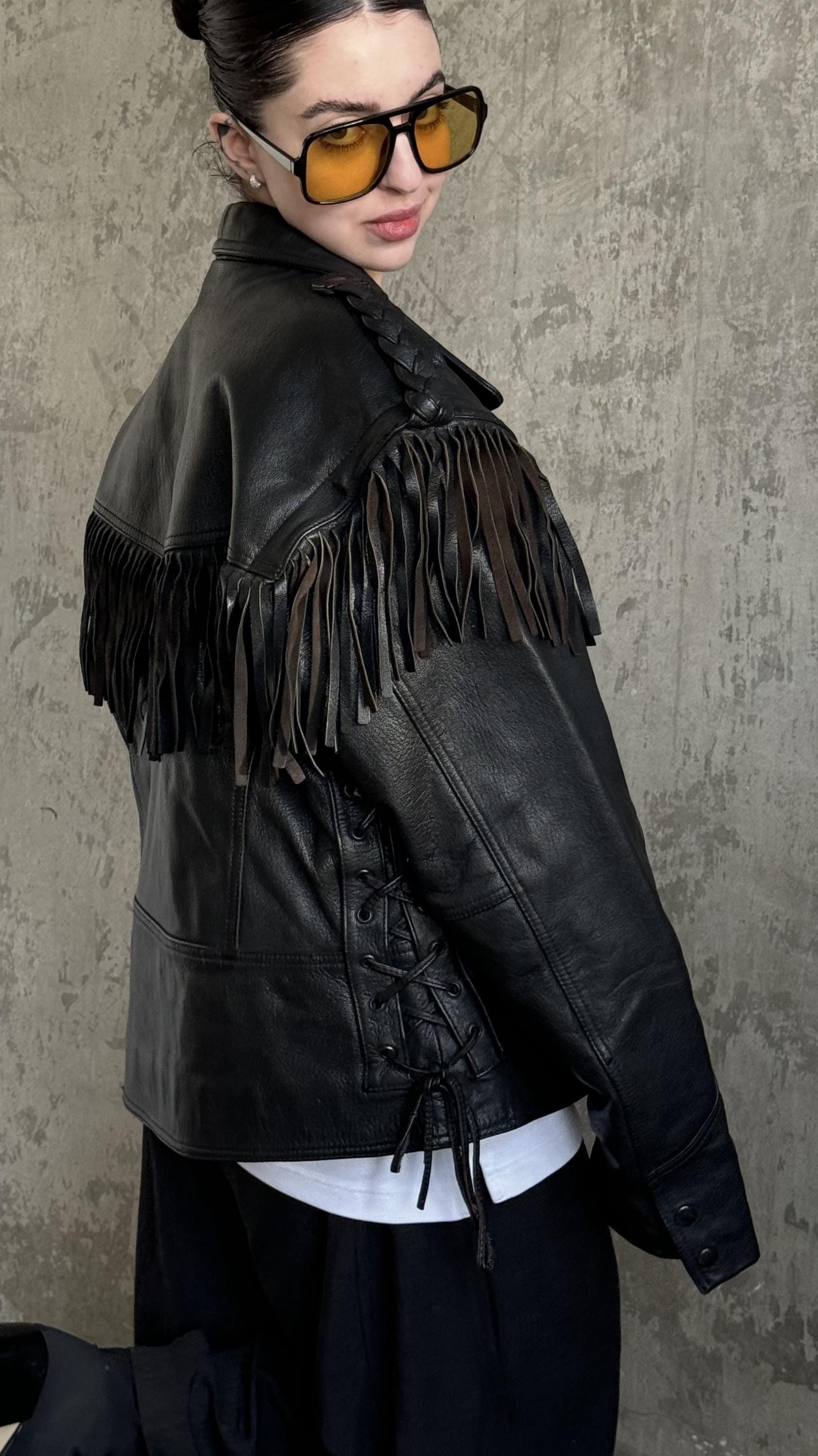 A woman wearing a black leather jacket with fringe, sunglasses, and a white shirt stands against a concrete wall.