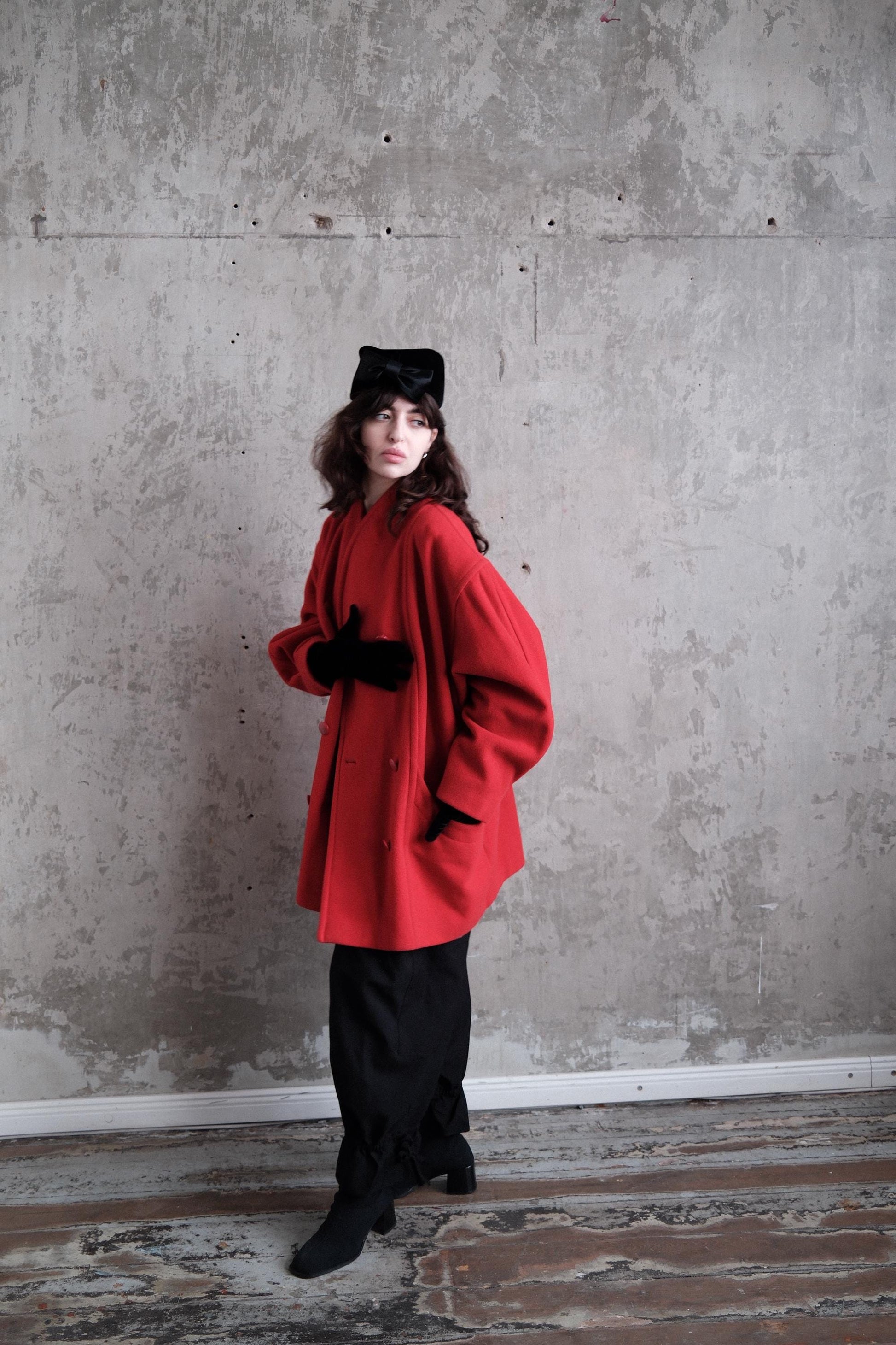 A person wearing a red coat and black pants stands in a room with a concrete wall.
