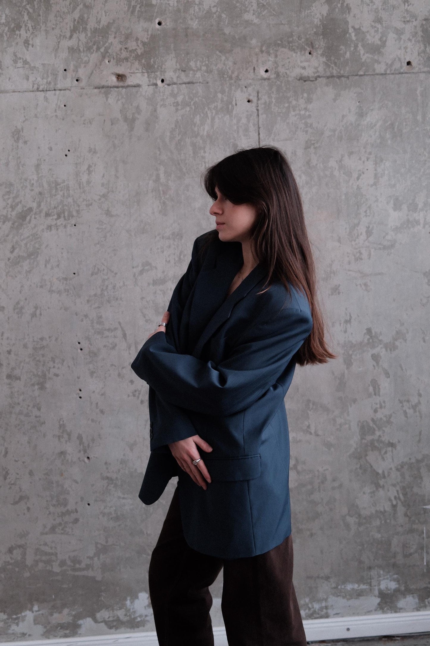 A woman in a dark jacket is leaning against a concrete wall, her long hair flowing freely.