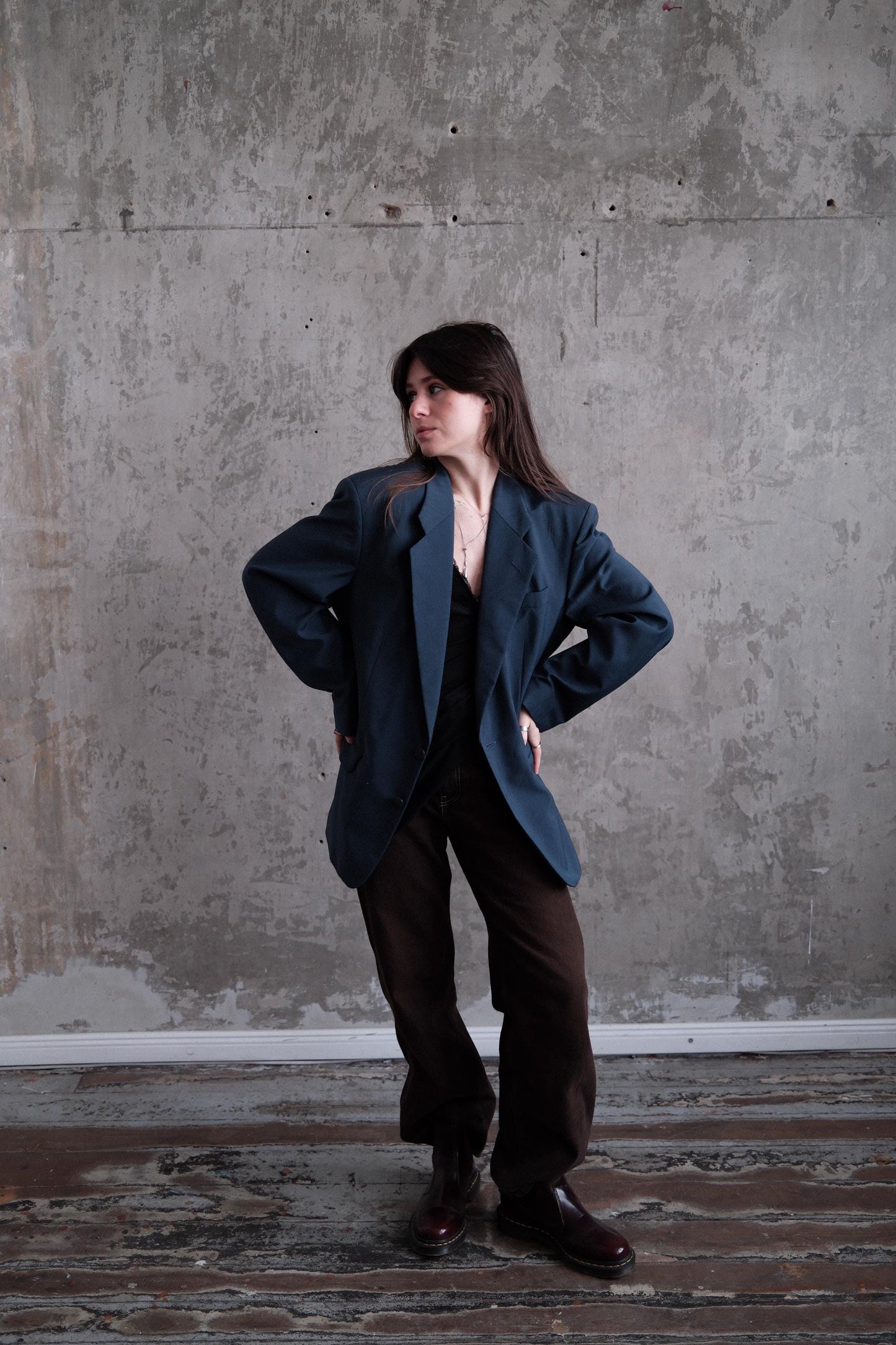 A person wearing a dark jacket and pants is standing against a concrete wall, with their arms crossed and leaning against the wall.