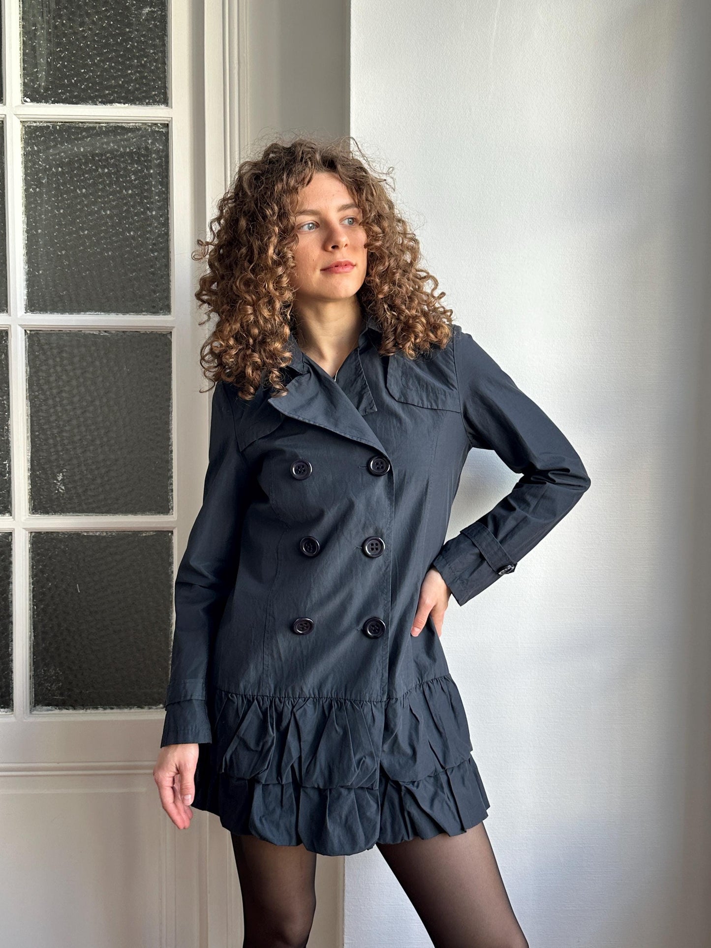 A woman with curly hair wearing a black coat and skirt is lying on her stomach in front of a window.