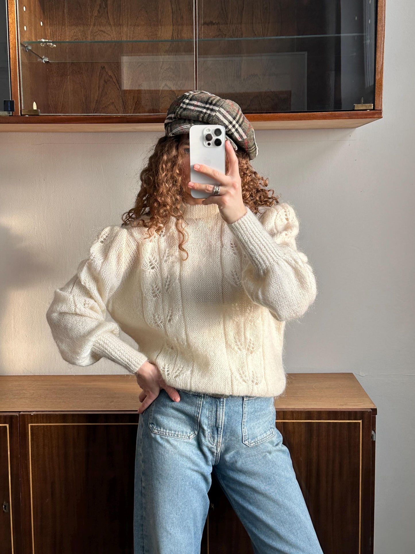 A person is taking a selfie with a white iPhone, wearing a white sweater, a plaid hat, and blue jeans.
