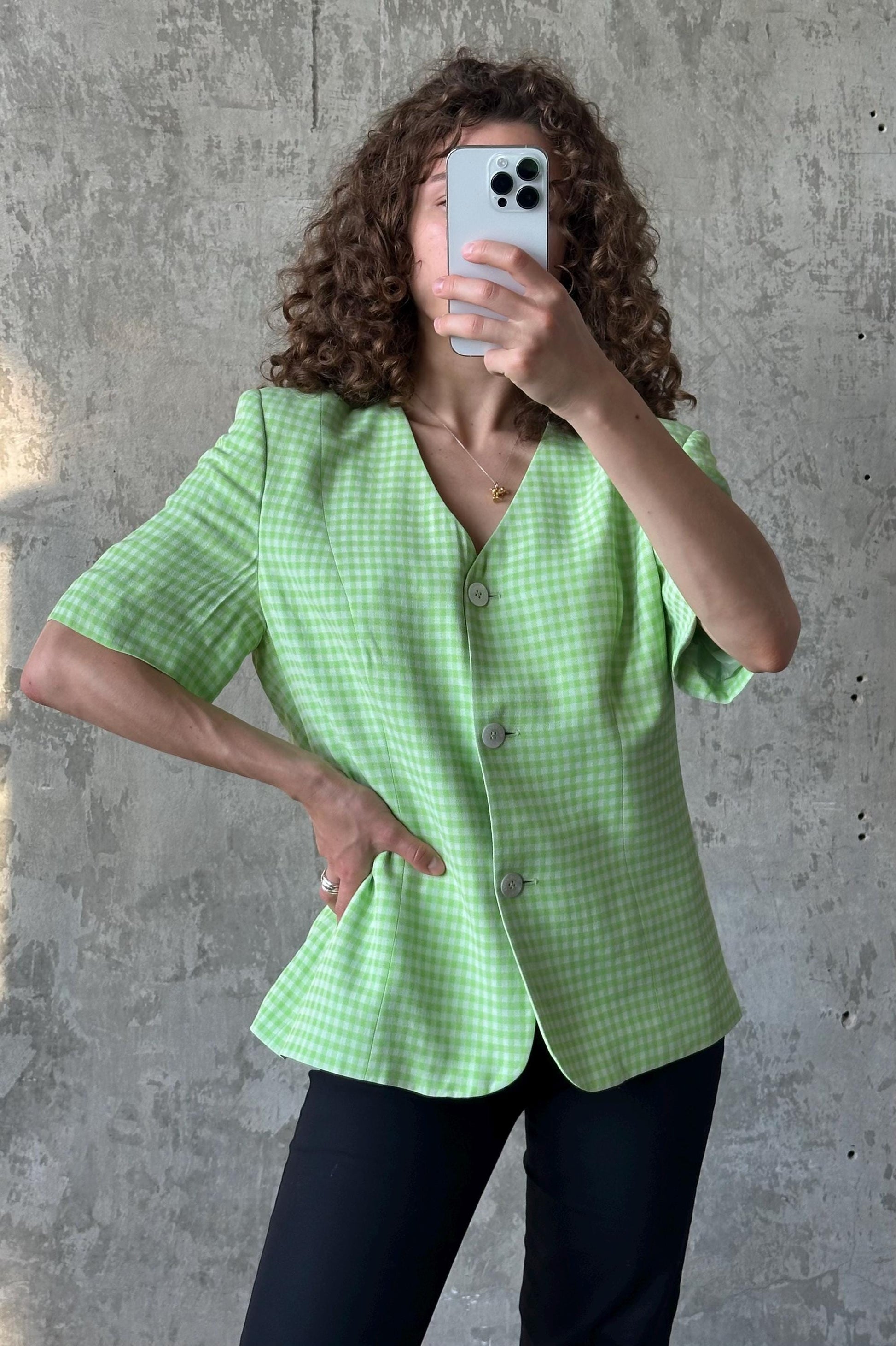 A woman with curly hair is taking a selfie with her phone, wearing a green and white checkered blazer.