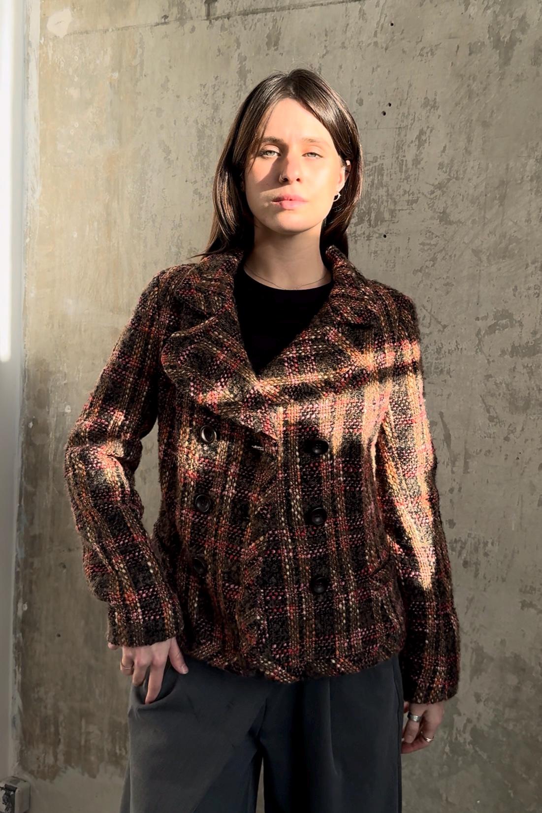 A woman wearing a brown and black plaid jacket stands in front of a concrete wall, looking directly at the camera with a serious expression.