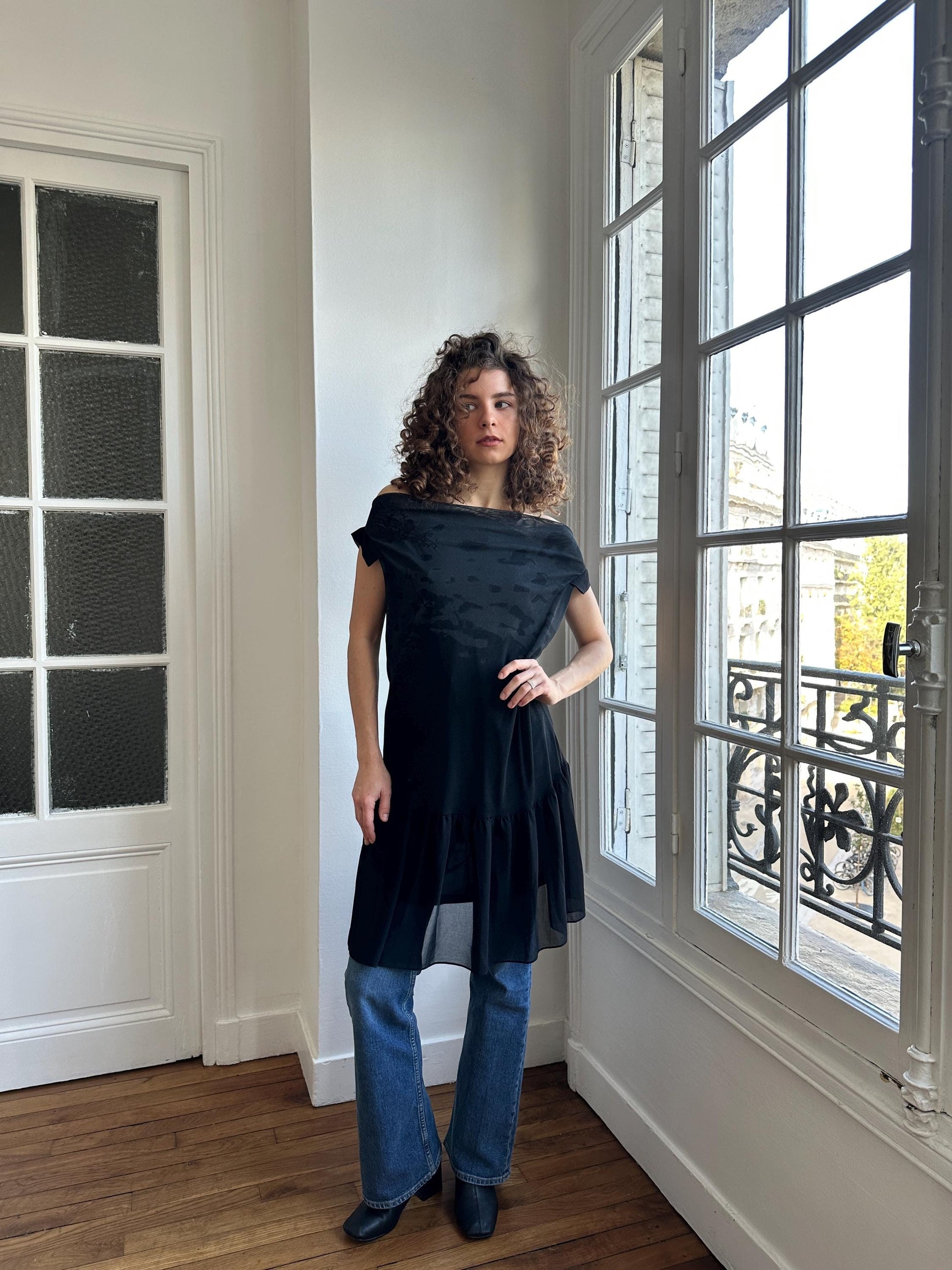 A woman with curly hair stands in a room, wearing a black off-the-shoulder dress and blue jeans.