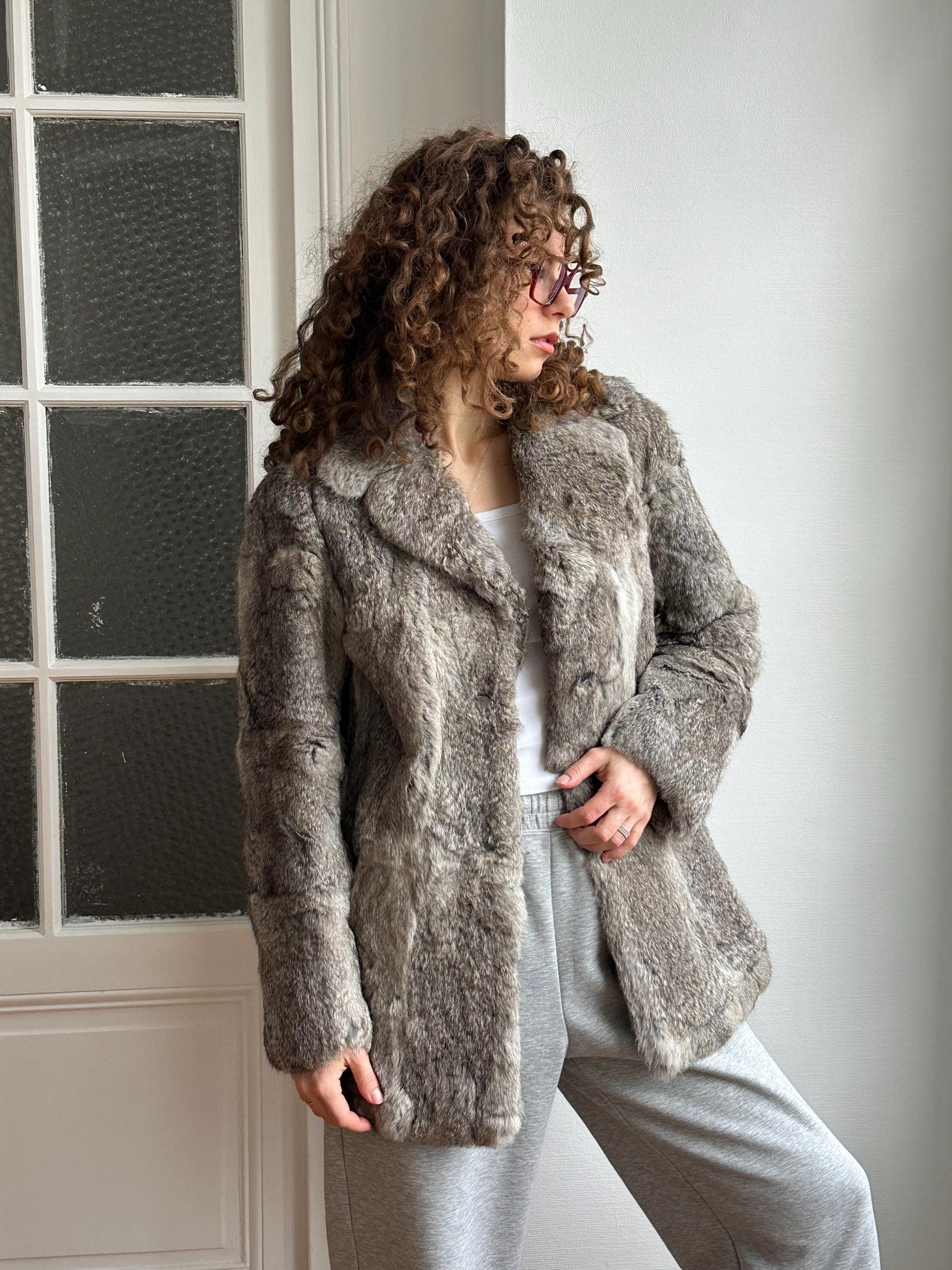 A woman with curly hair wearing a gray fur coat and glasses stands in front of a window.