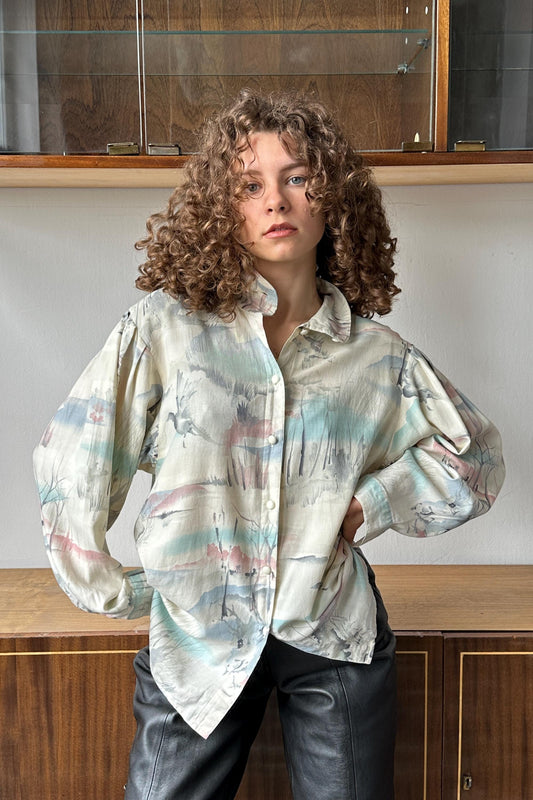 A young woman with curly hair wearing a white blouse with a colorful print stands in front of a wooden cabinet.