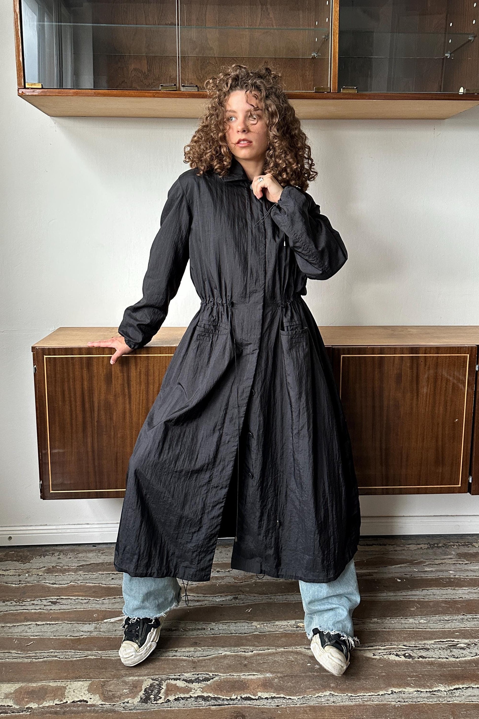 A woman with curly hair wearing a long black coat stands in a room with wooden cabinets and a wooden floor.