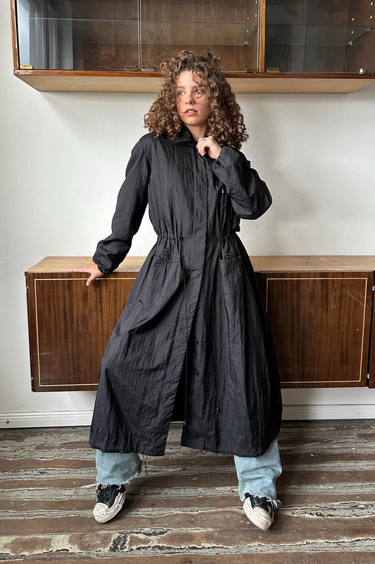 A woman with curly hair wearing a long black coat stands in a room with wooden cabinets and a wooden floor.