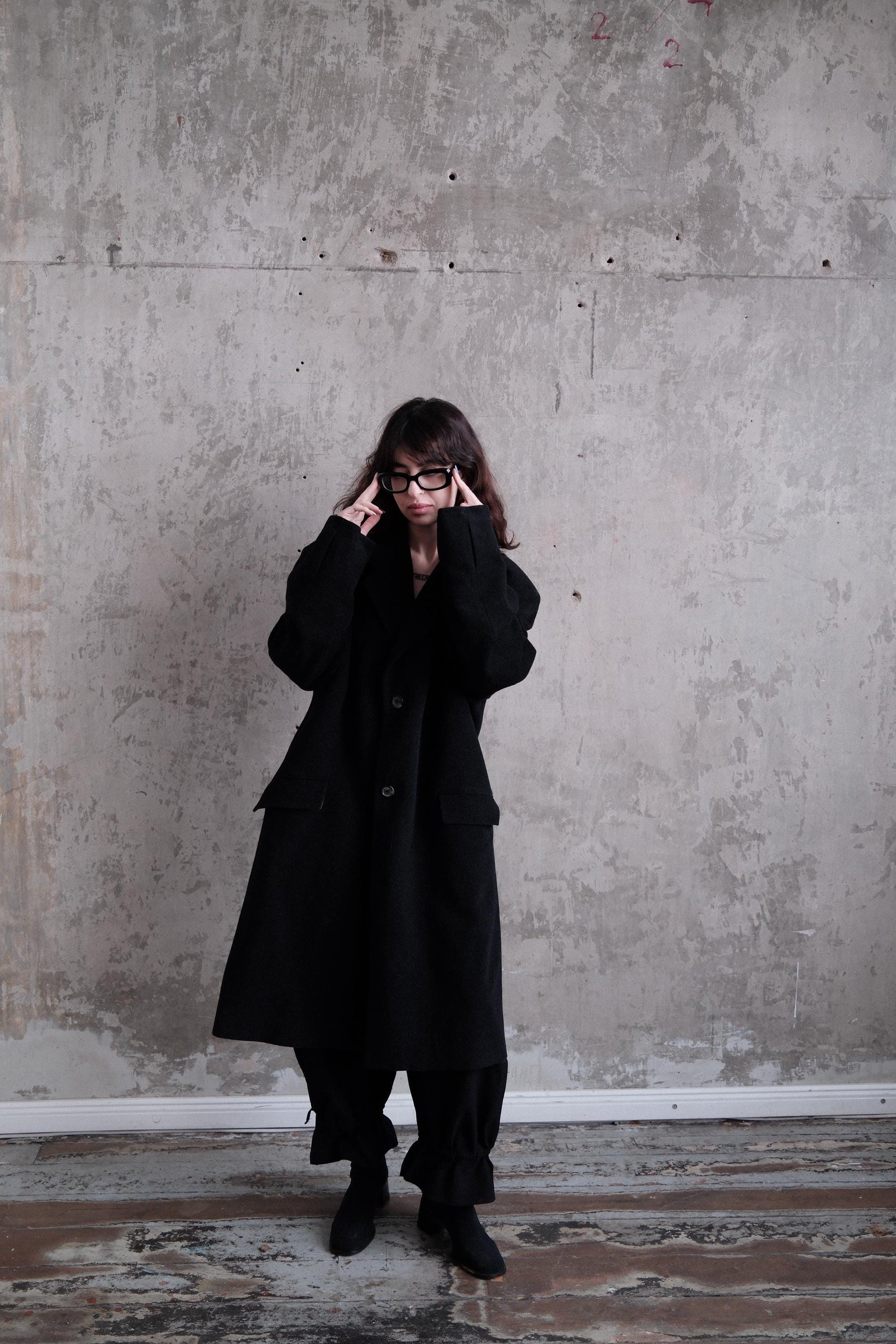 A person wearing a long black coat stands in a room with a concrete wall.