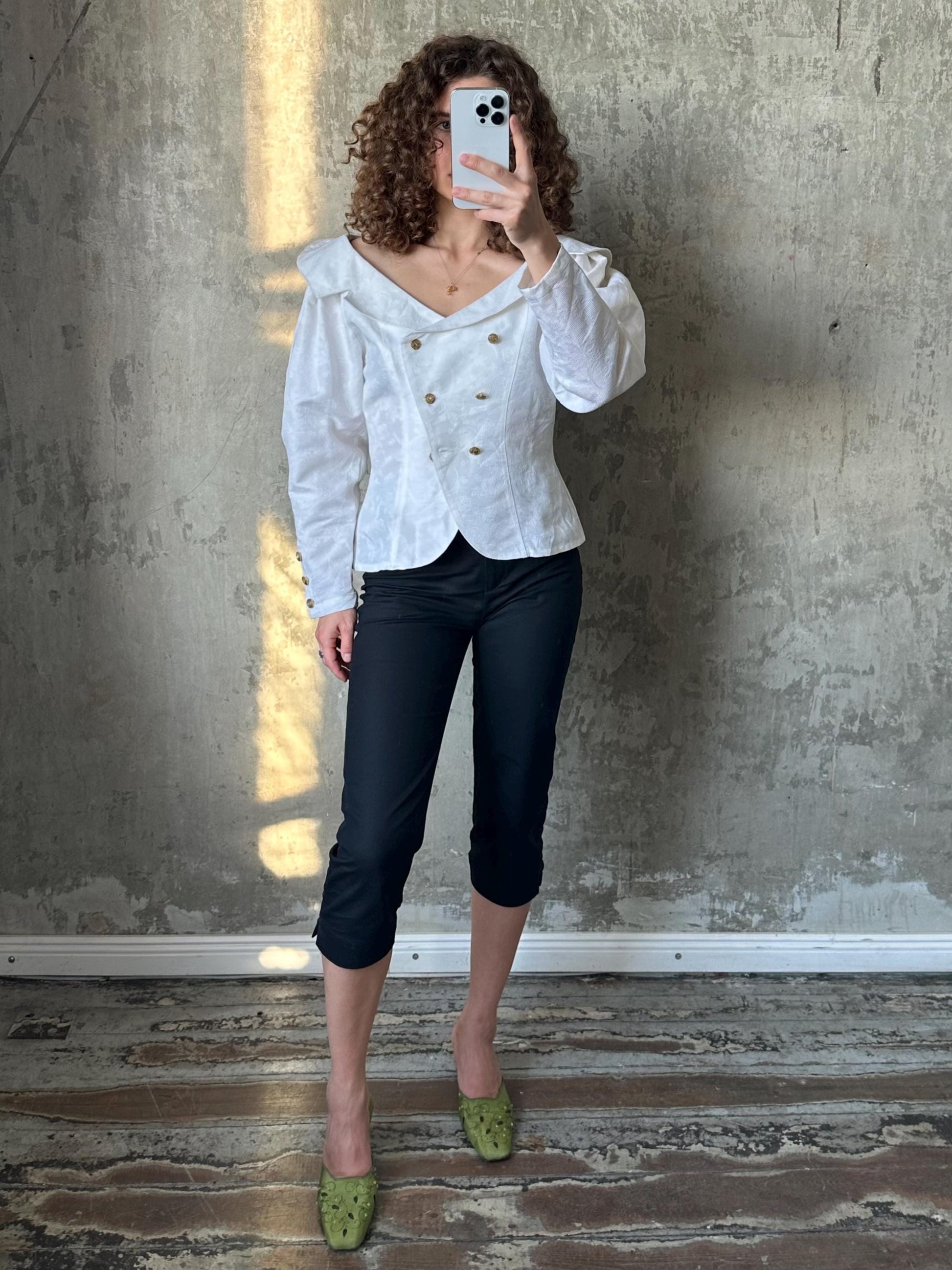 A woman with curly hair is taking a selfie in front of a concrete wall, wearing a white blouse, black pants, and green shoes.