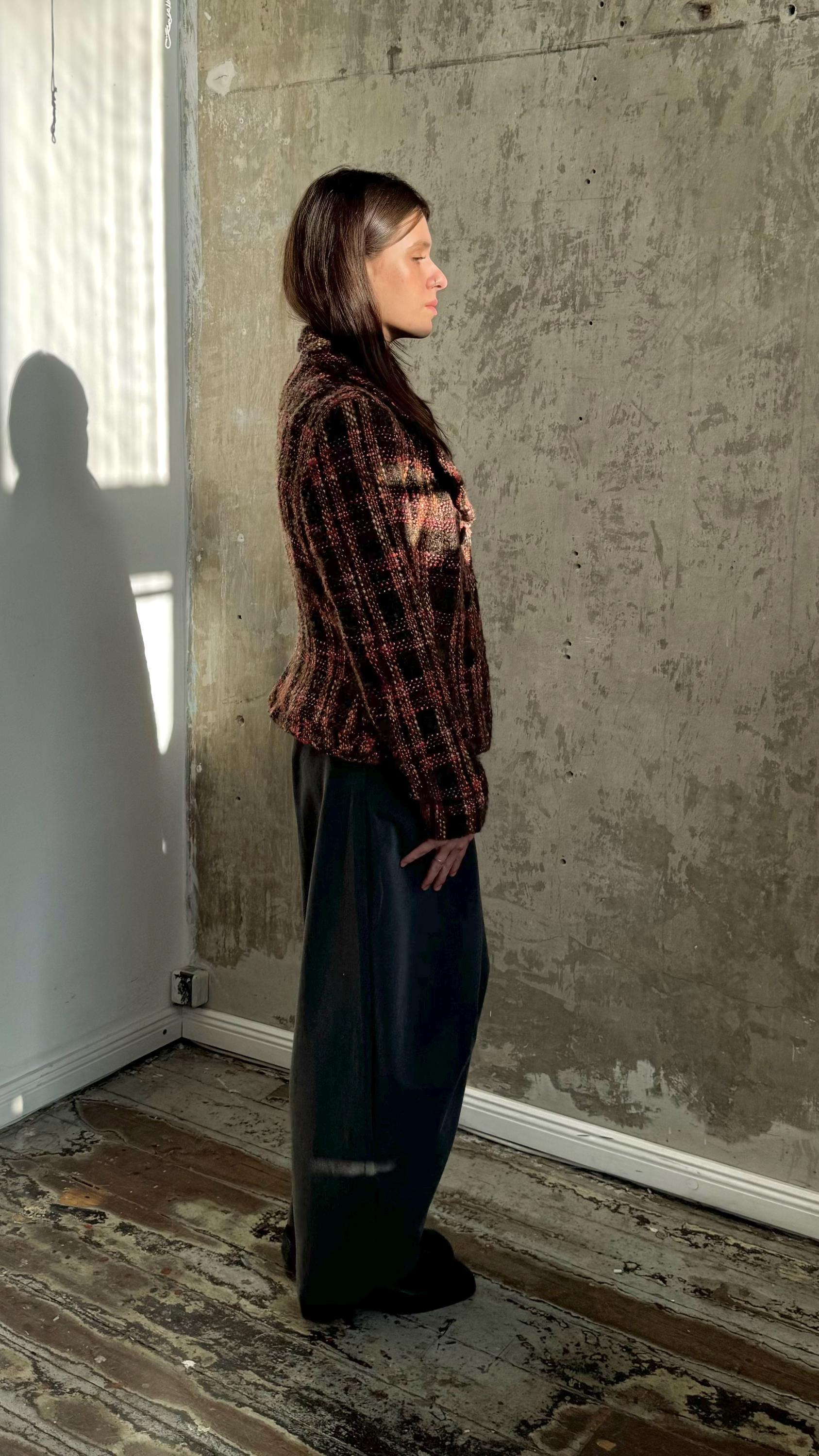 A woman wearing a plaid jacket stands in a room with a concrete wall and wooden floor.