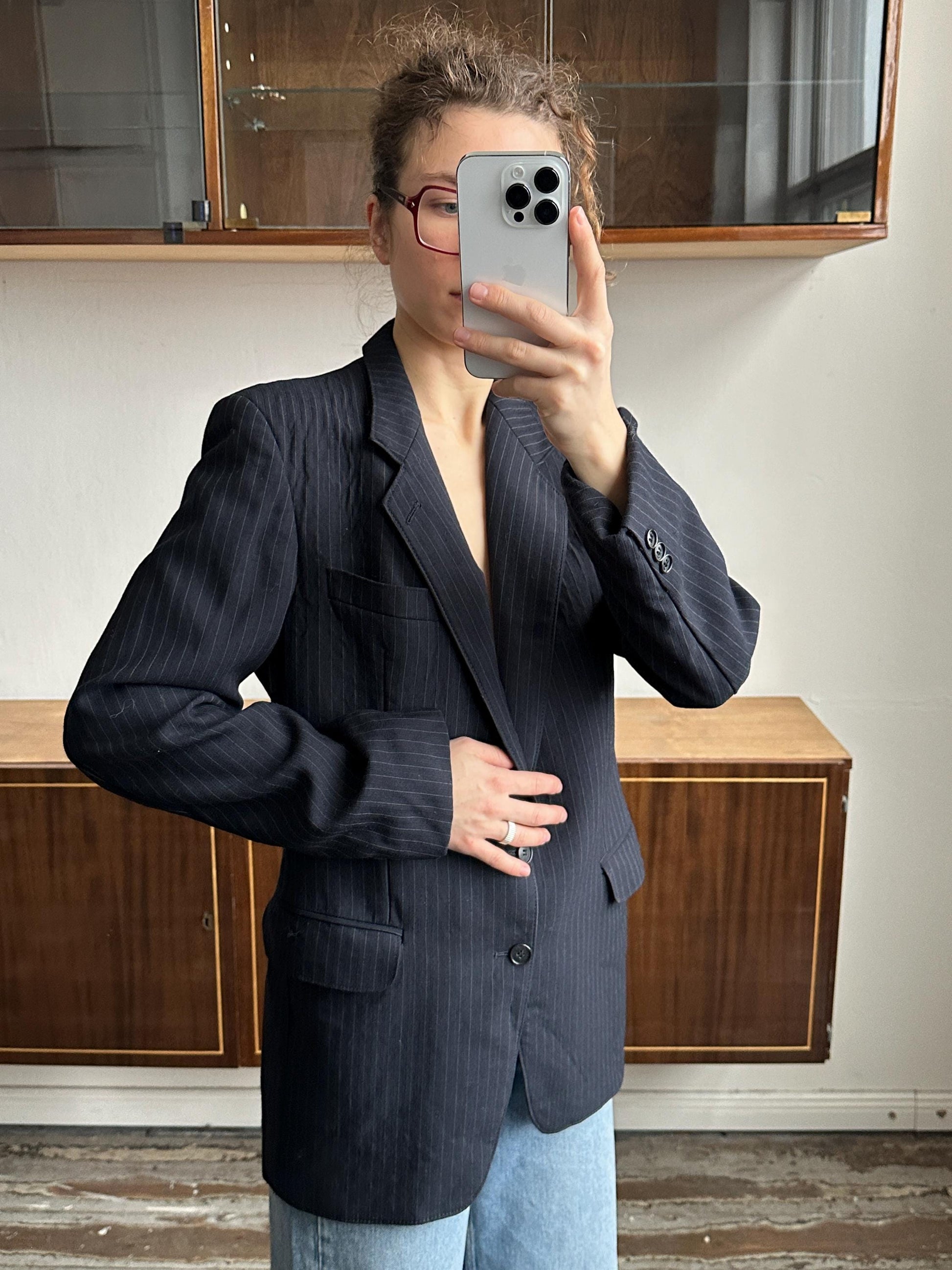 A person is taking a selfie with a smartphone, wearing a black blazer and blue jeans, in a room with wooden furniture.