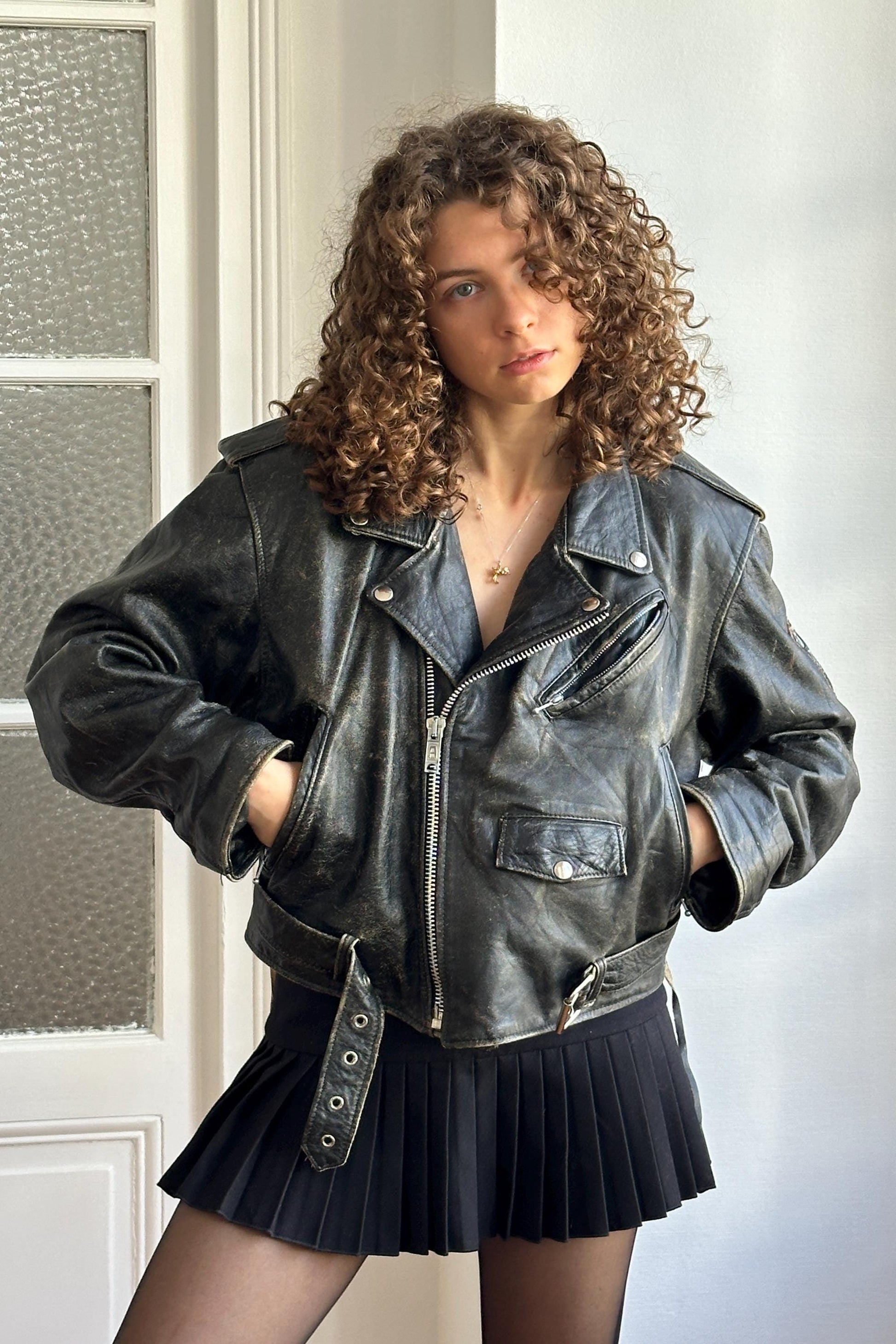 A young woman with curly hair wearing a black leather jacket and a black pleated skirt stands in front of a white door.