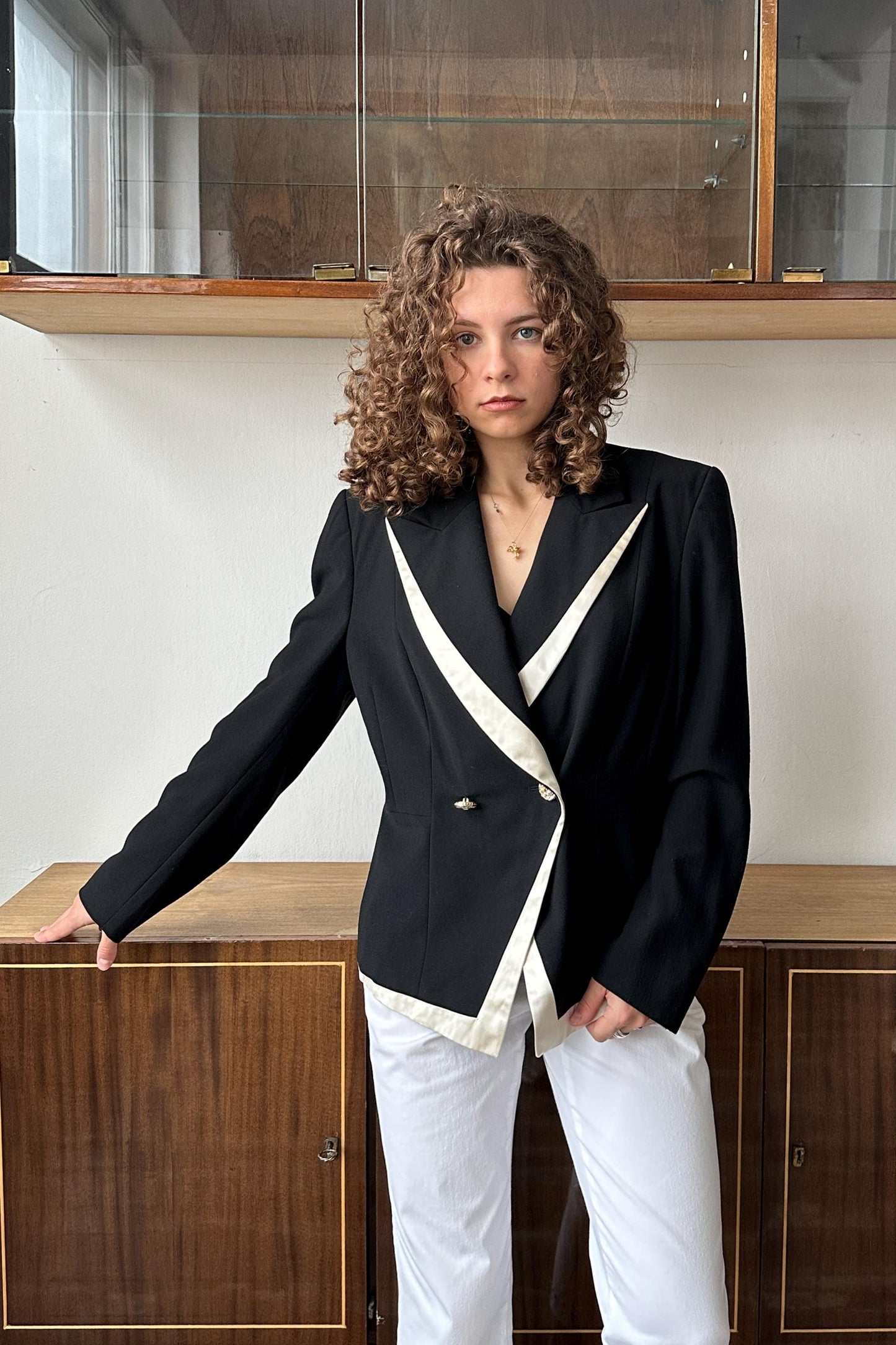 A woman with curly hair wearing a black and white blazer stands in front of a wooden cabinet.