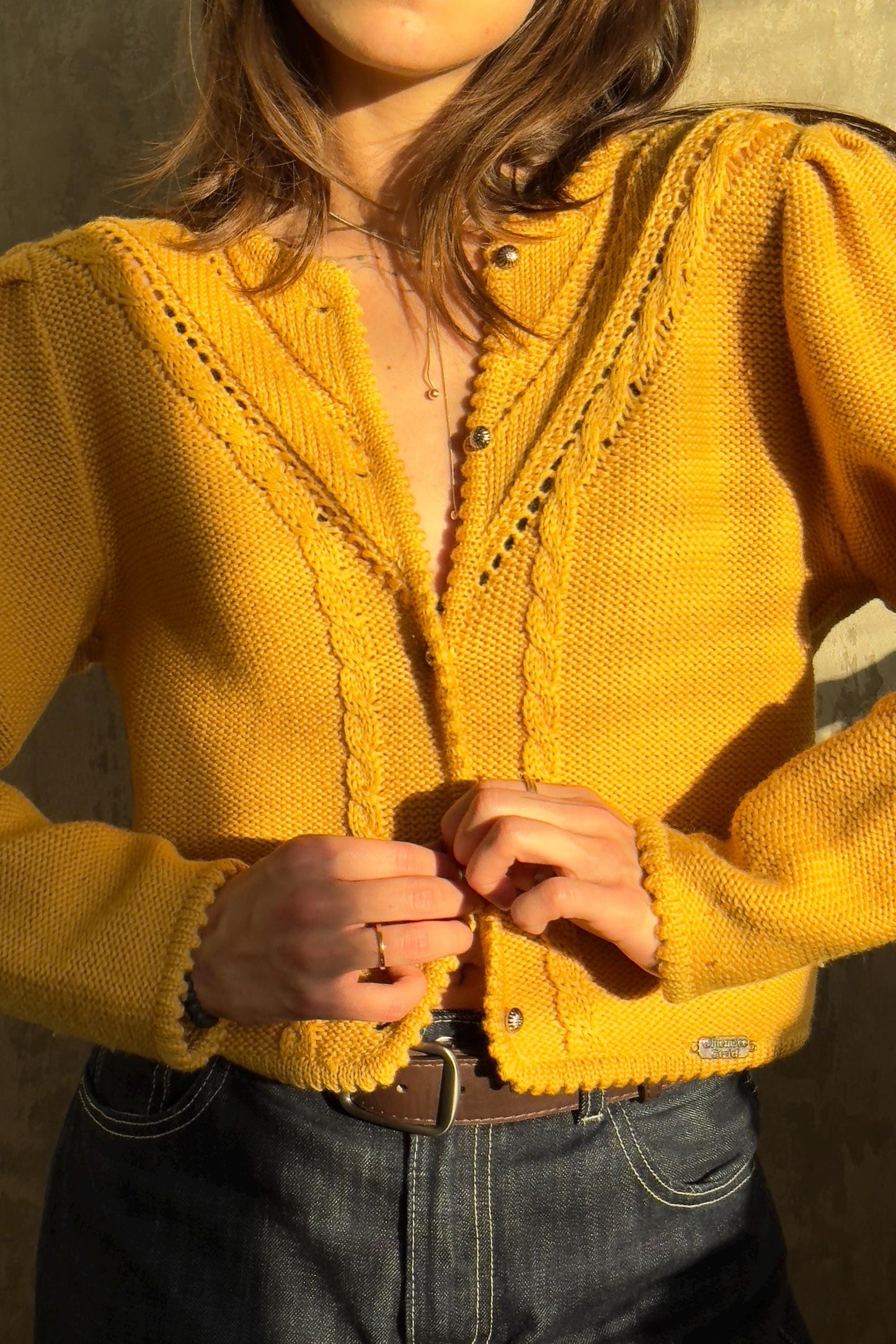 A woman wearing a yellow cardigan and jeans stands against a concrete wall, adjusting the buttons of her sweater.
