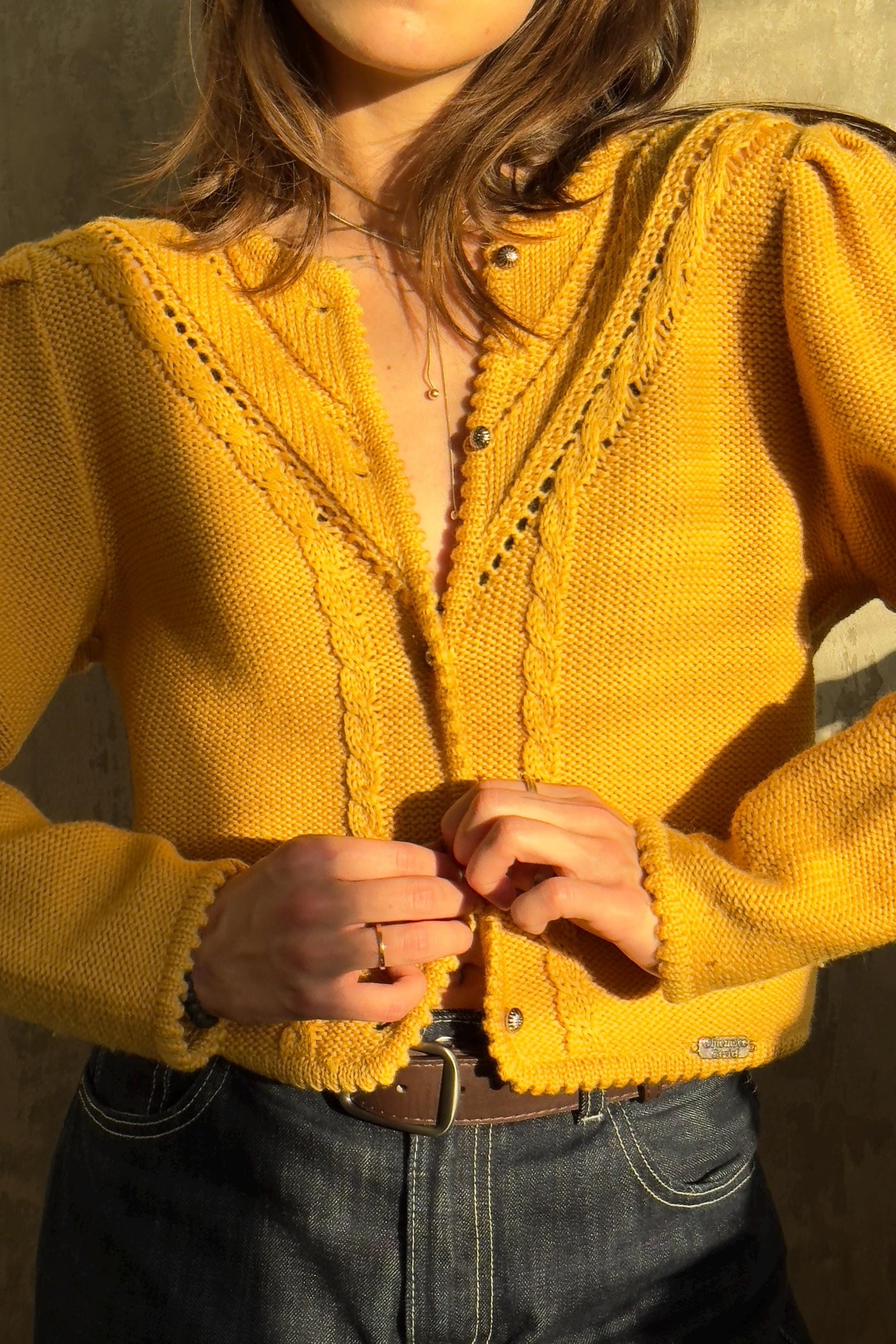 A woman wearing a yellow cardigan and jeans stands against a concrete wall, adjusting the buttons of her sweater.