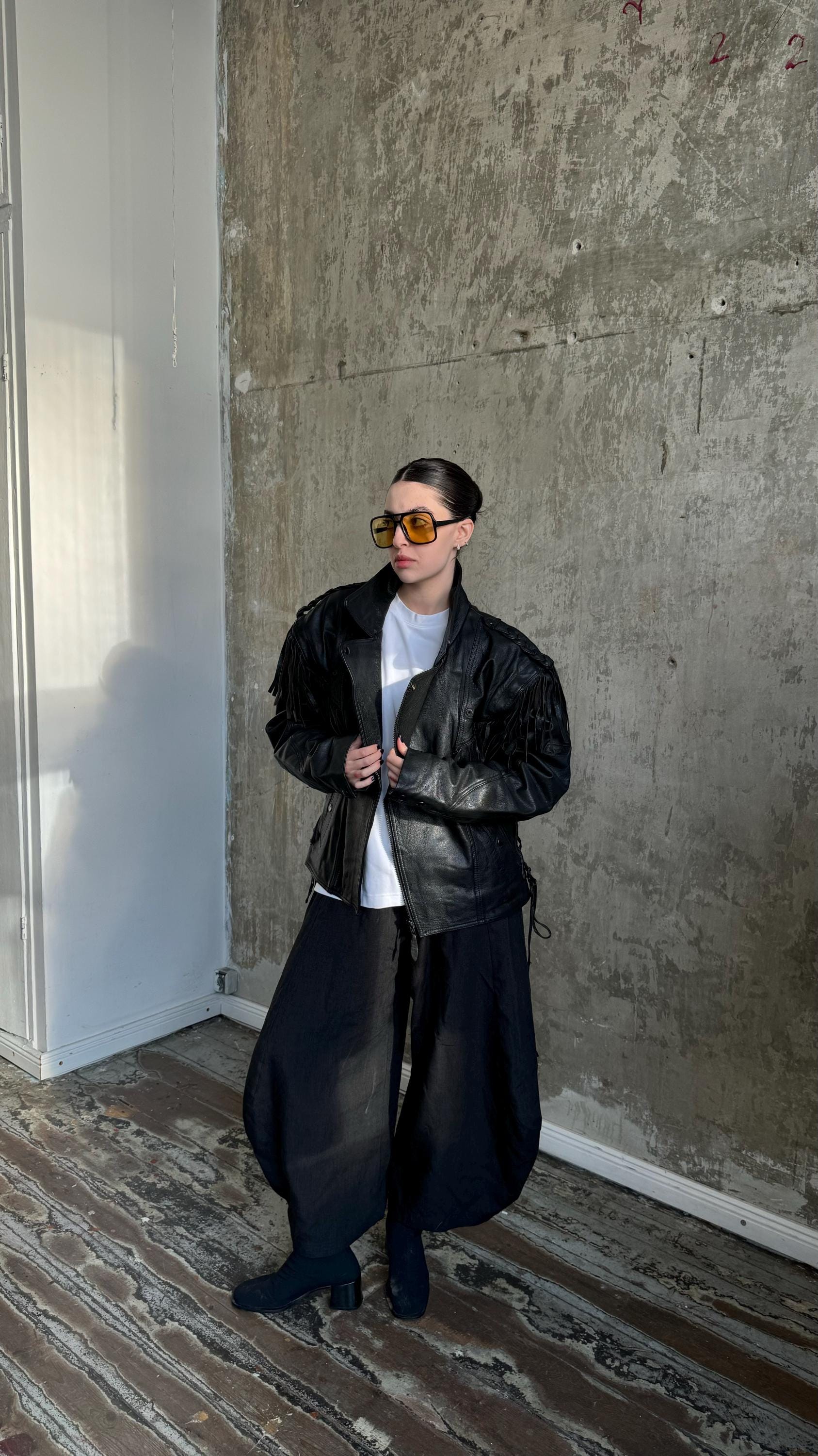 A person wearing a black leather jacket, sunglasses, and a white shirt stands in front of a concrete wall.