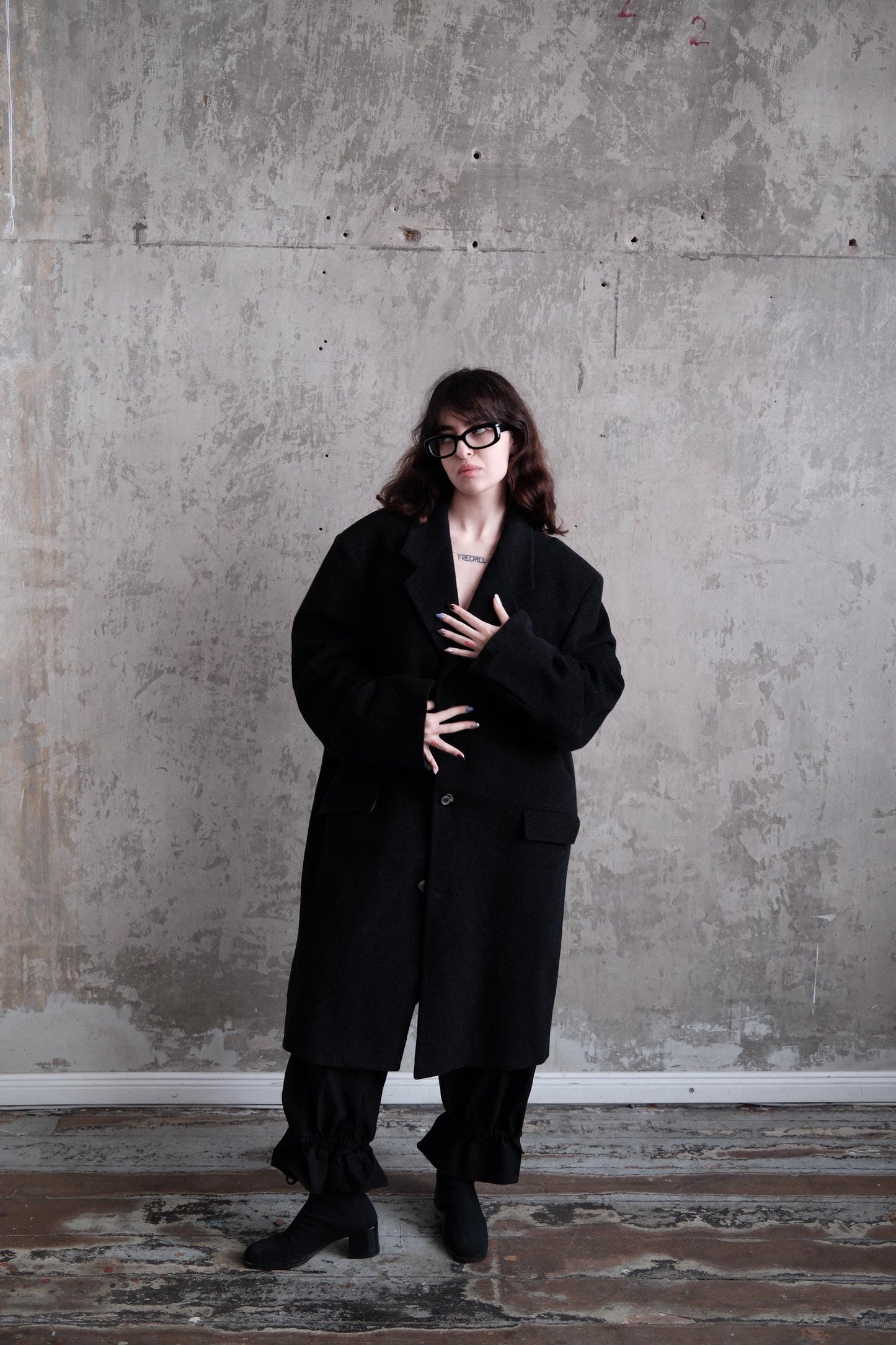 A person wearing a long black coat stands in a room with a concrete wall.