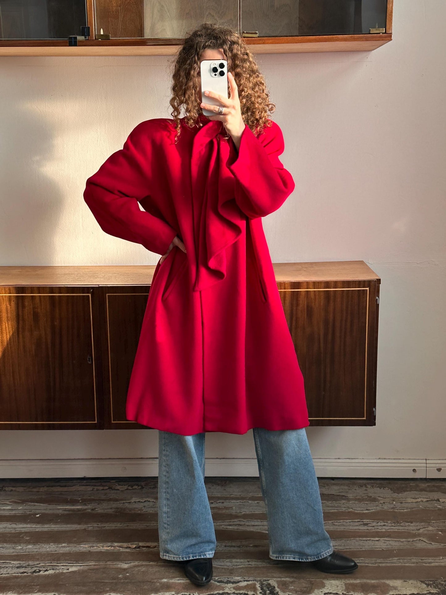 A person wearing a red coat and jeans is taking a selfie with a smartphone.