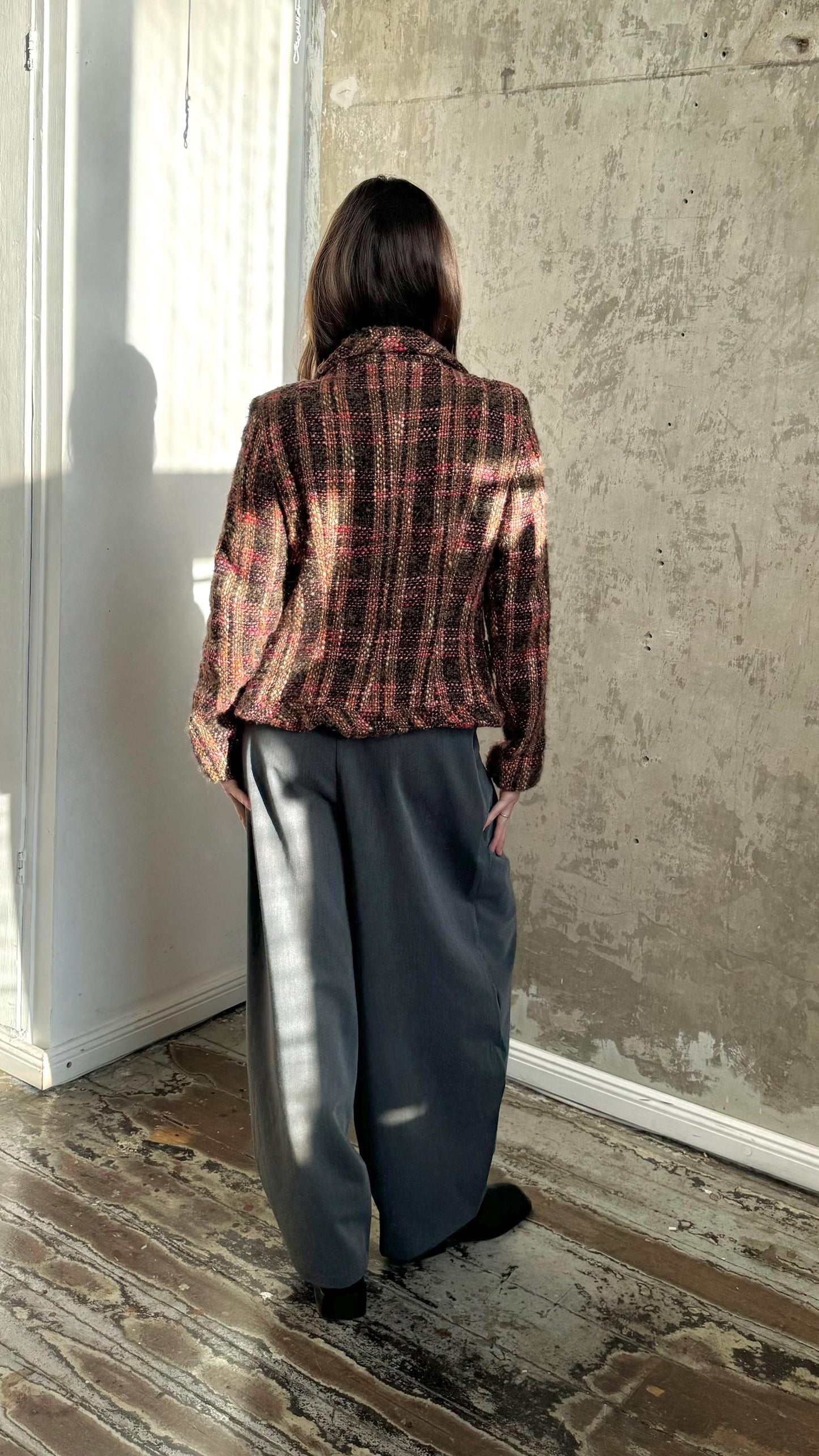 A person wearing a checkered jacket and wide-leg pants stands in front of a concrete wall, facing away from the camera.