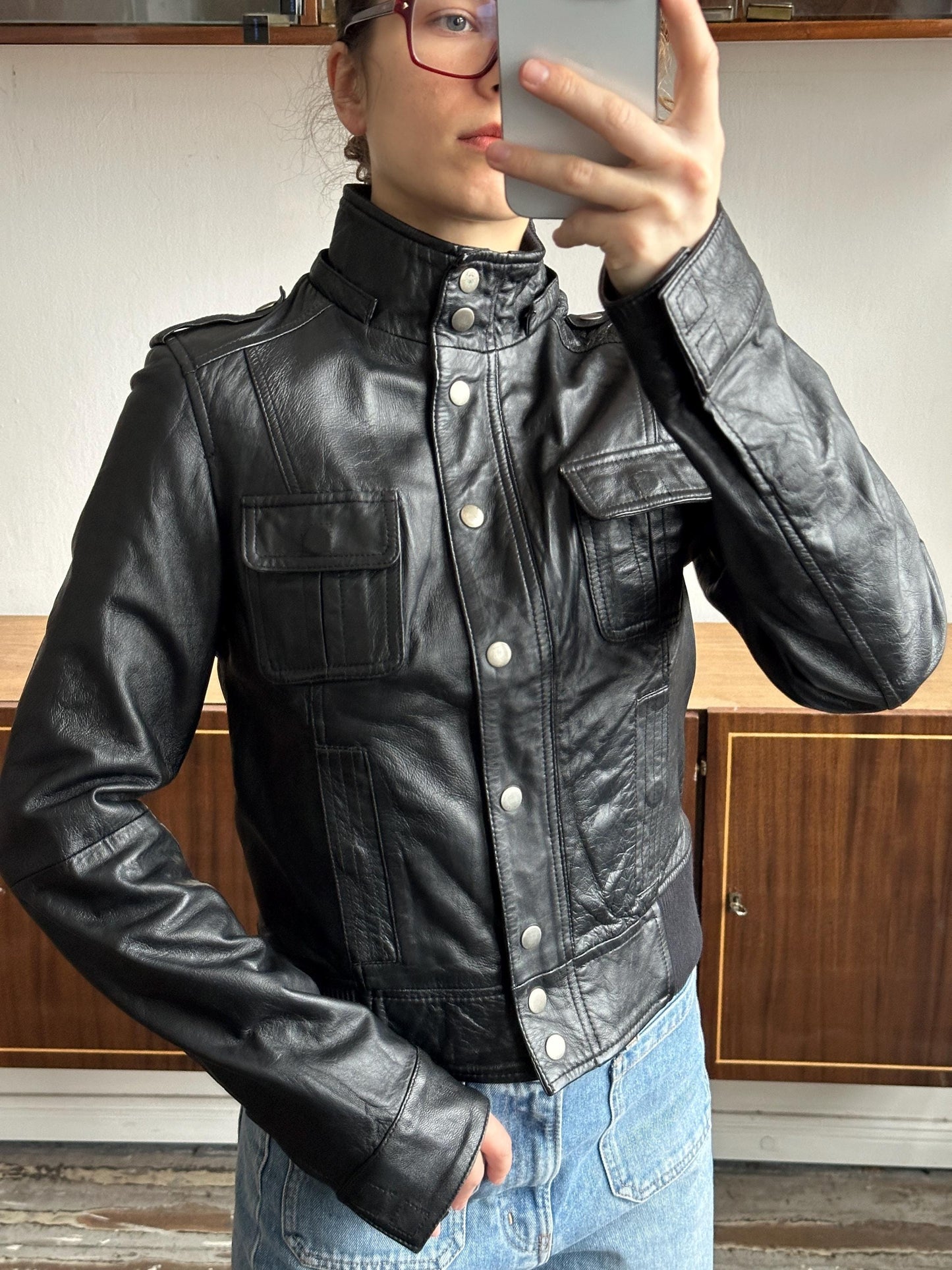 A person wearing a black leather jacket and jeans is taking a selfie with a smartphone.