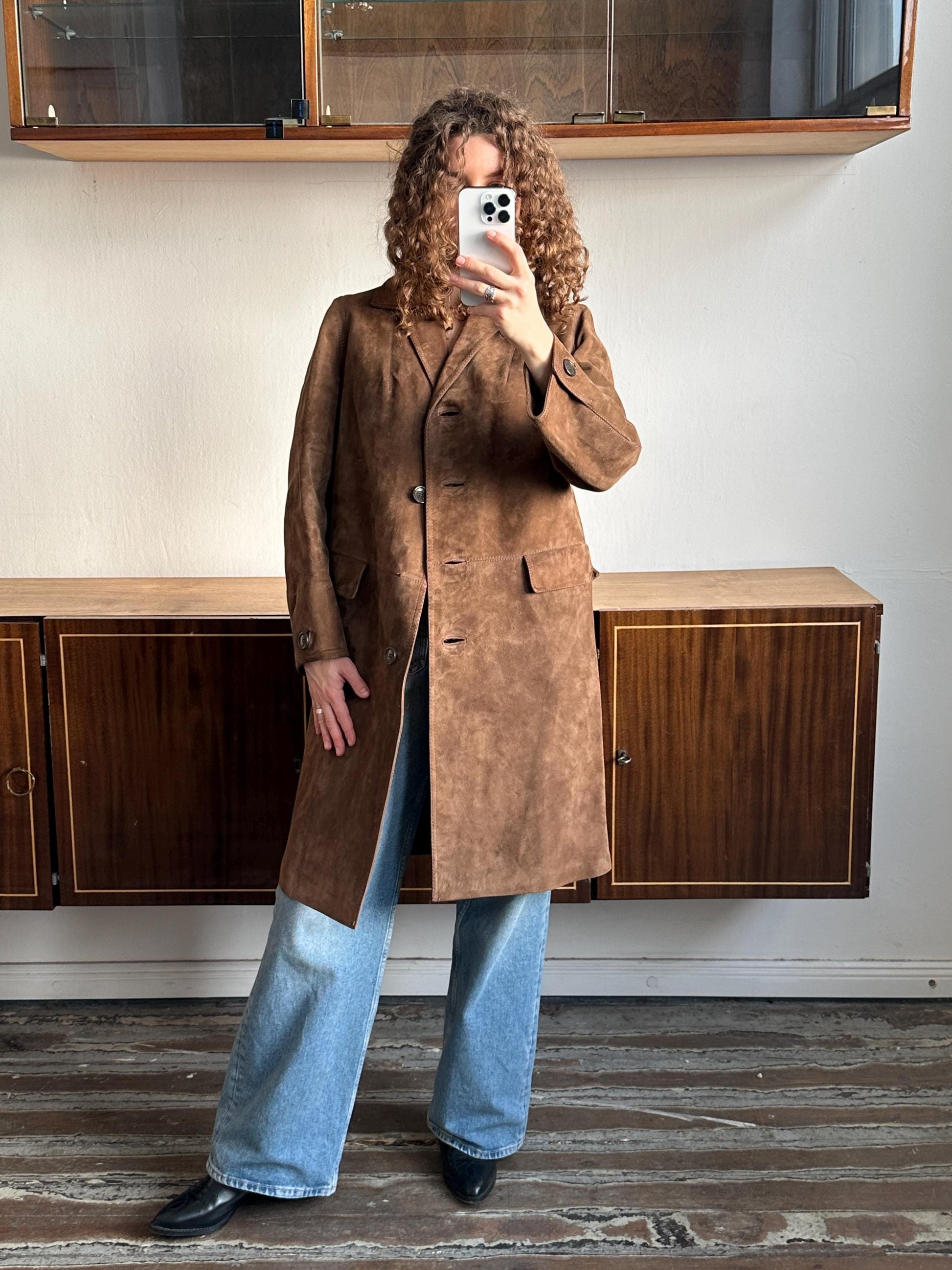 A person is taking a selfie with a brown coat and blue jeans, standing in front of a wooden cabinet.