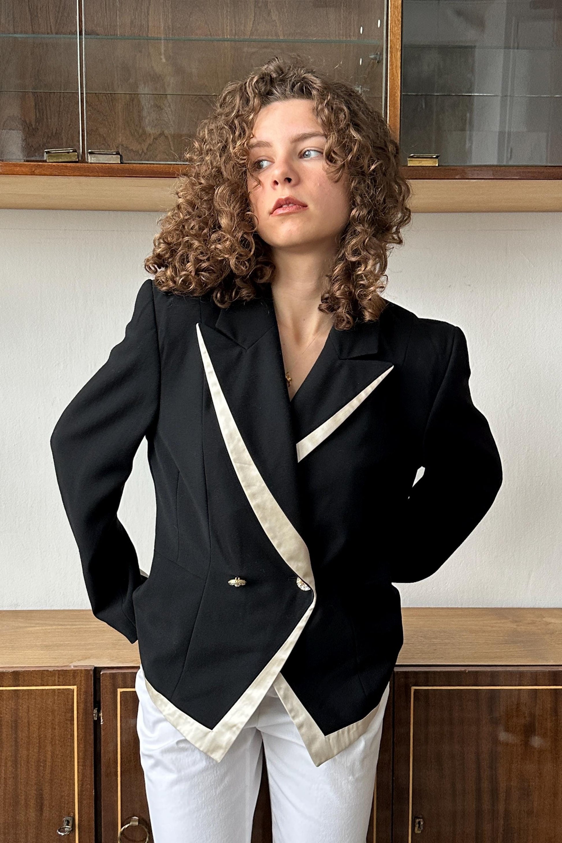 A woman with curly hair wearing a black blazer with white accents stands in front of a wooden cabinet.