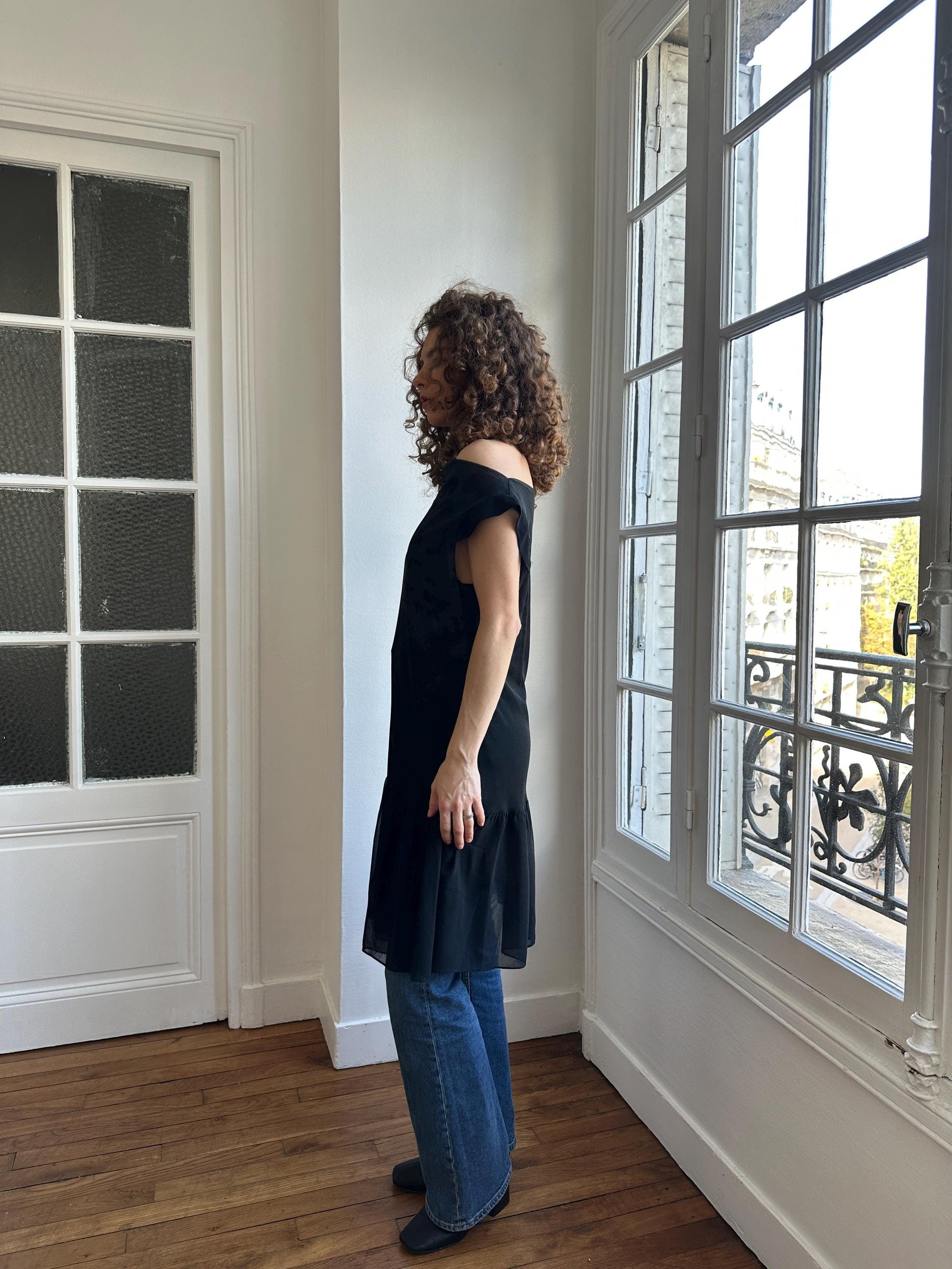 A woman with curly hair stands in a room with a large window, wearing a black dress and blue jeans.