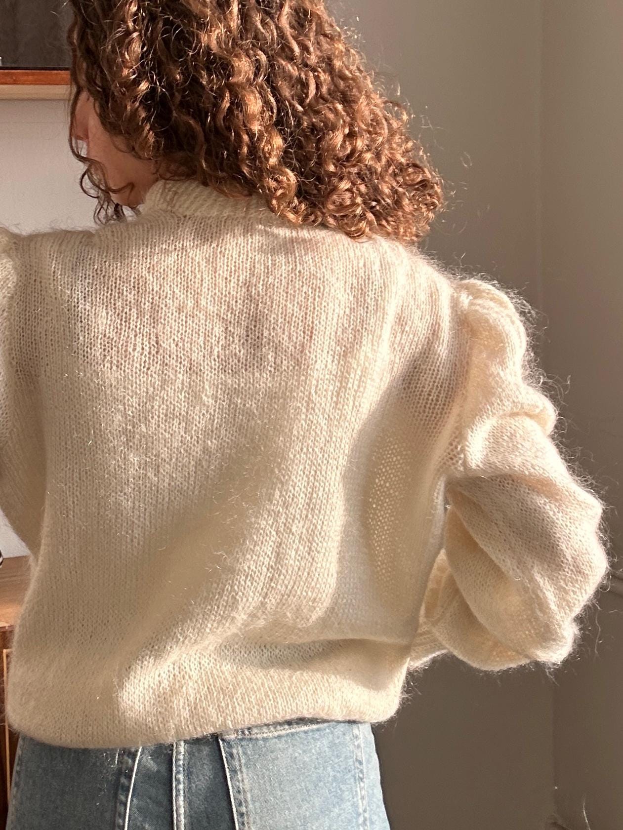 a person with curly hair wearing a white sweater and blue jeans, with their back facing the camera.
