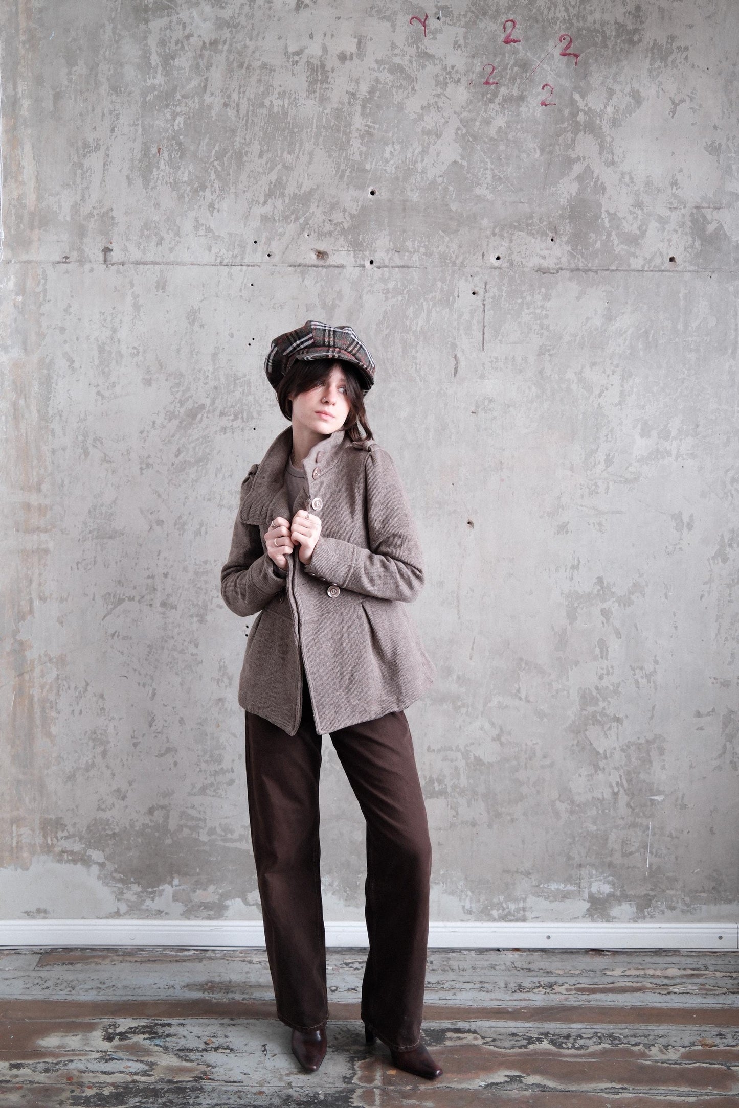 A person wearing a brown coat and hat stands in a room with a concrete wall and a wooden floor.