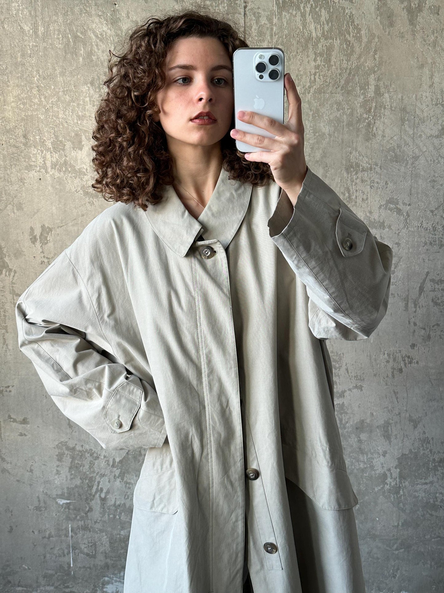 A woman with curly hair is taking a selfie with a smartphone, wearing a beige coat.