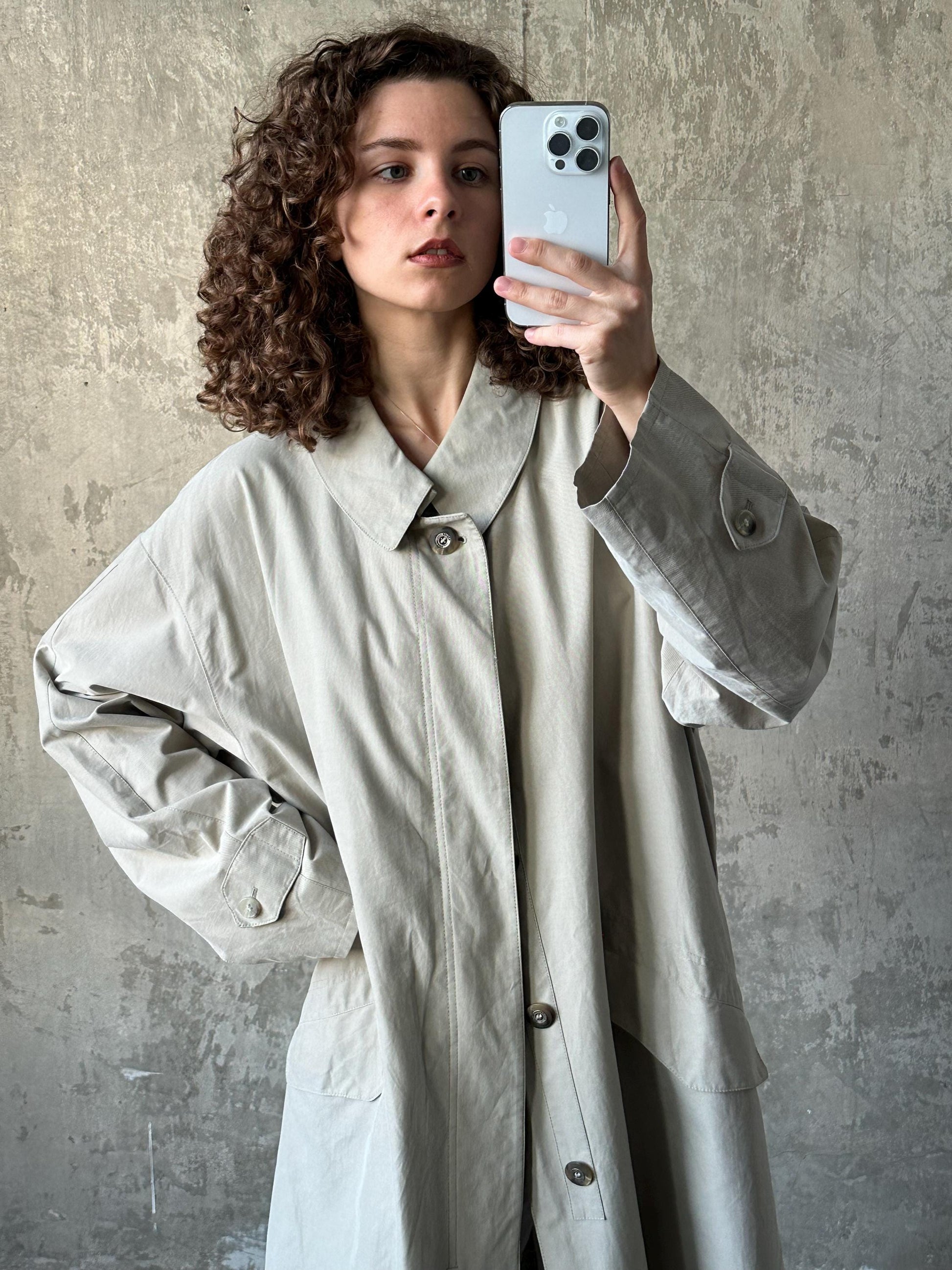A woman with curly hair is taking a selfie with a smartphone, wearing a beige coat.