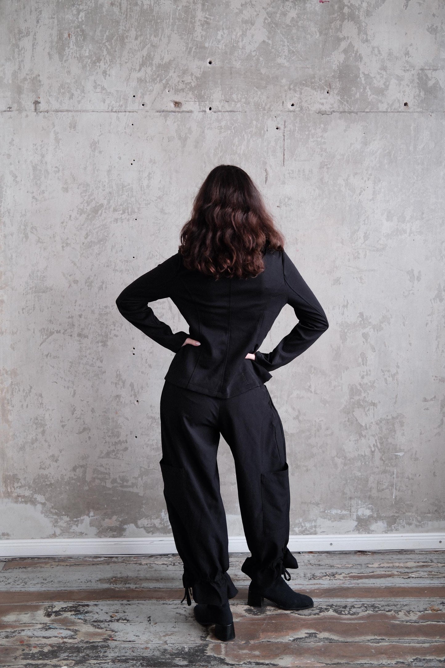A person in a black outfit stands with their hands on their hips, facing away from the camera, against a plain concrete wall.