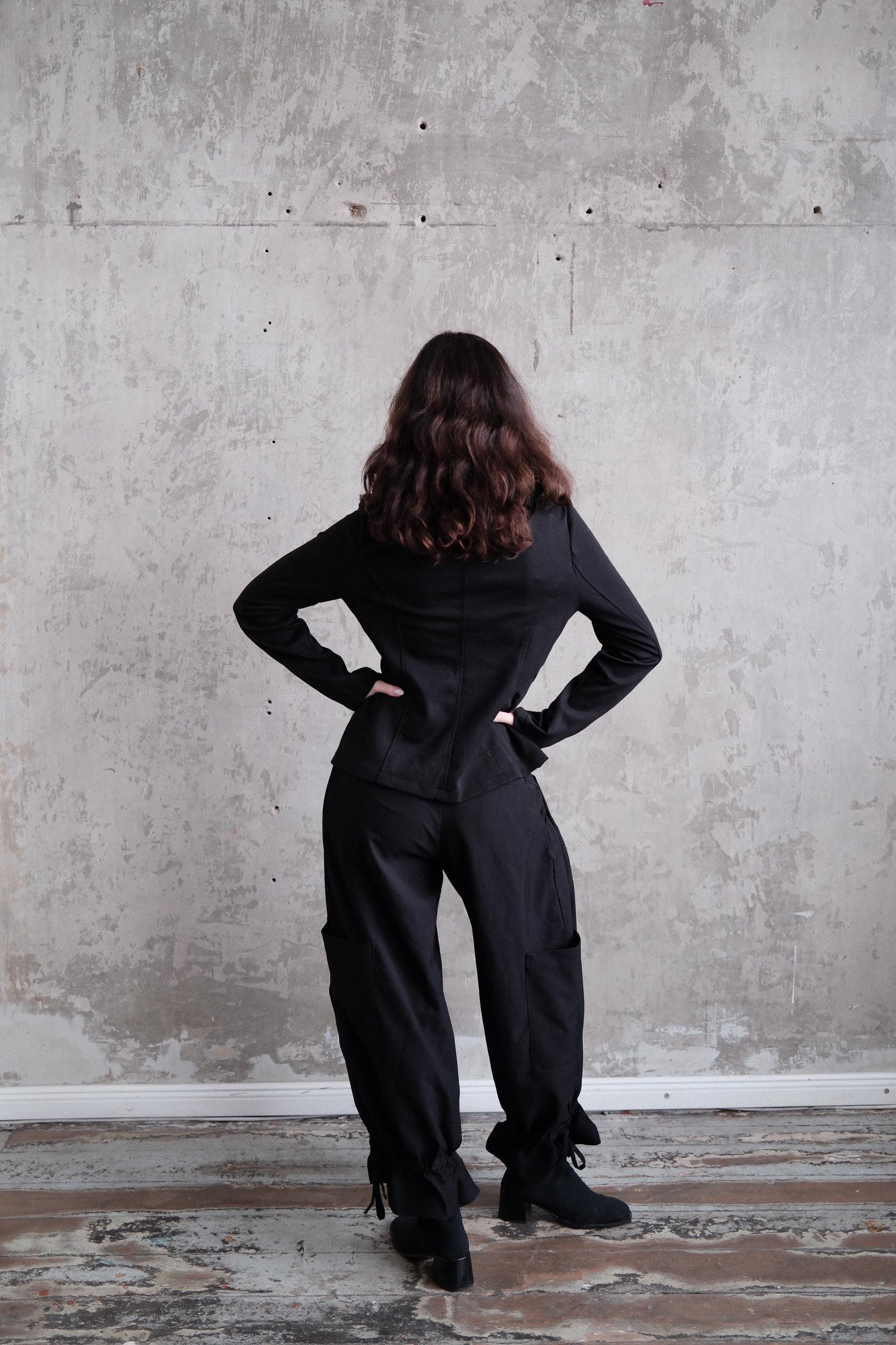 A person in a black outfit stands with their hands on their hips, facing away from the camera, against a plain concrete wall.