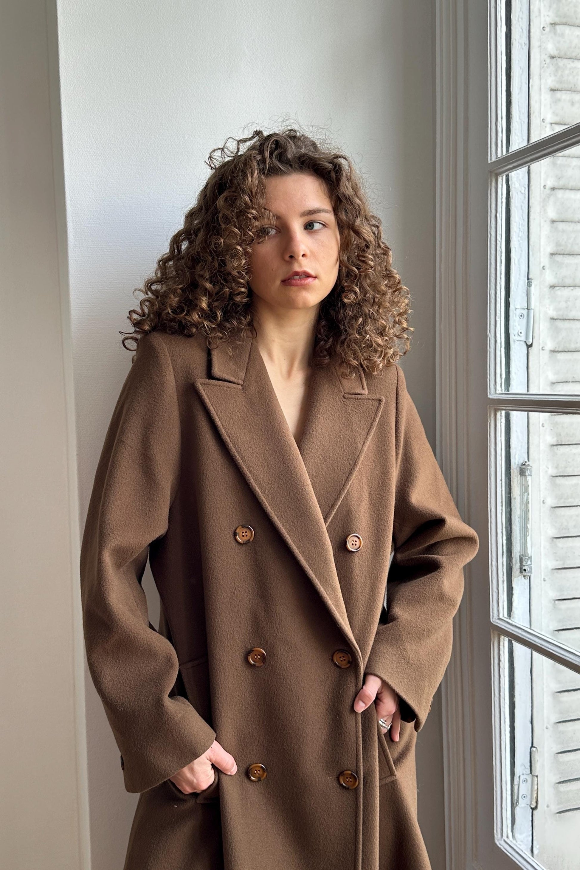 A woman with curly hair wearing a brown coat stands in front of a window, looking off to the side with a serious expression.