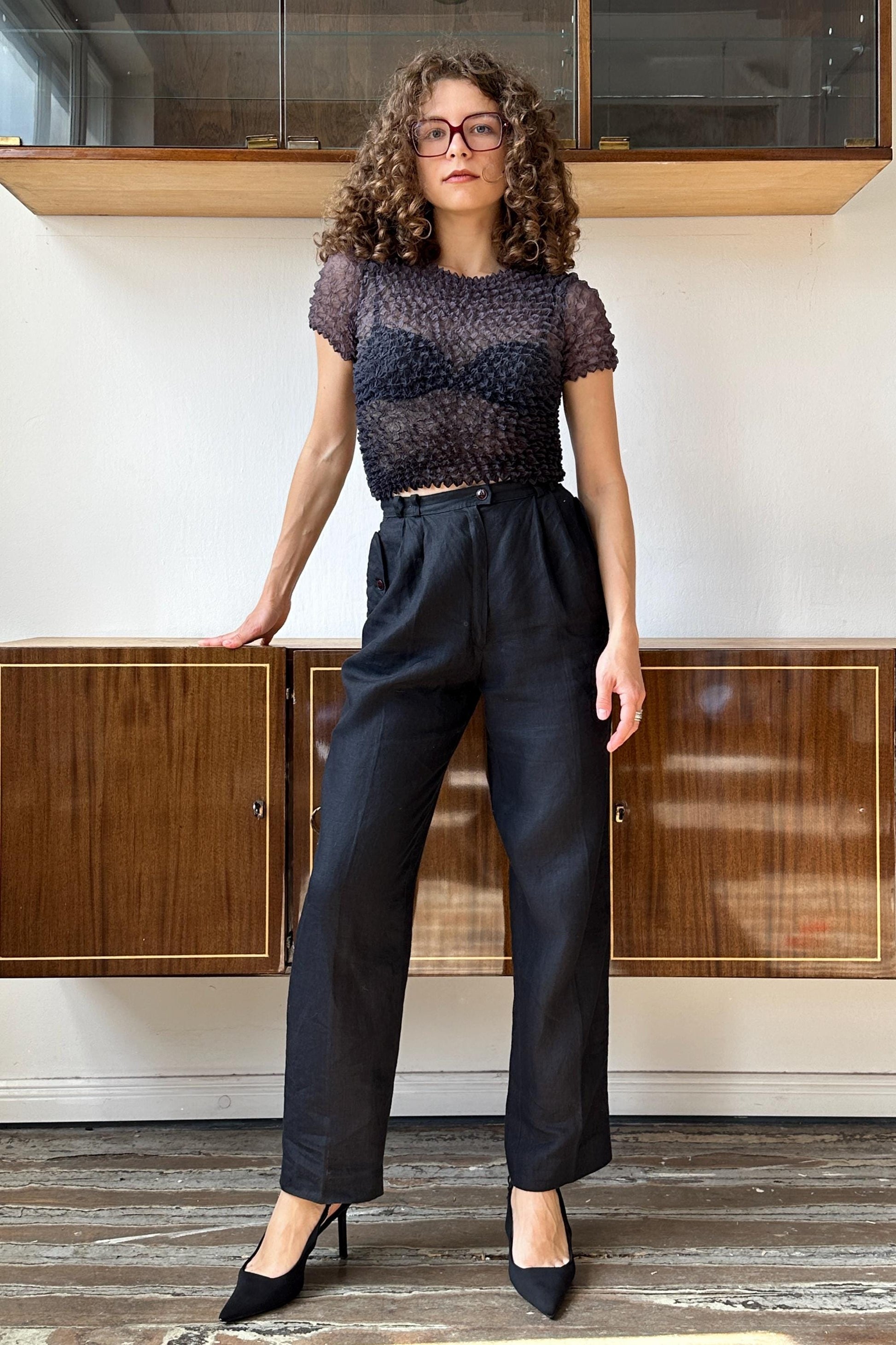 A woman with curly hair and glasses is standing in front of a wooden cabinet, wearing a black top and black pants.