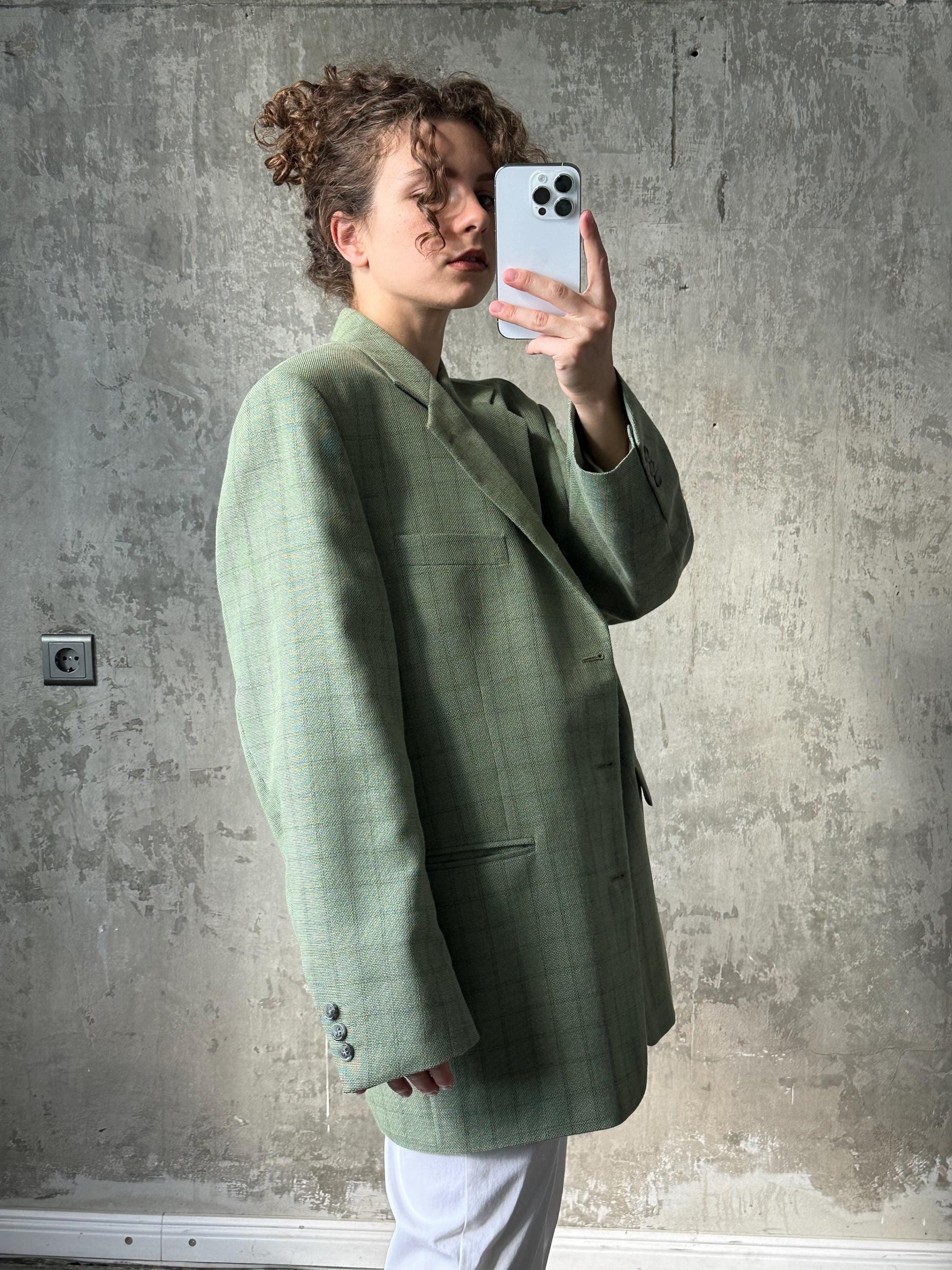 A woman in a green plaid blazer is taking a selfie with a smartphone, capturing her reflection in the mirror.
