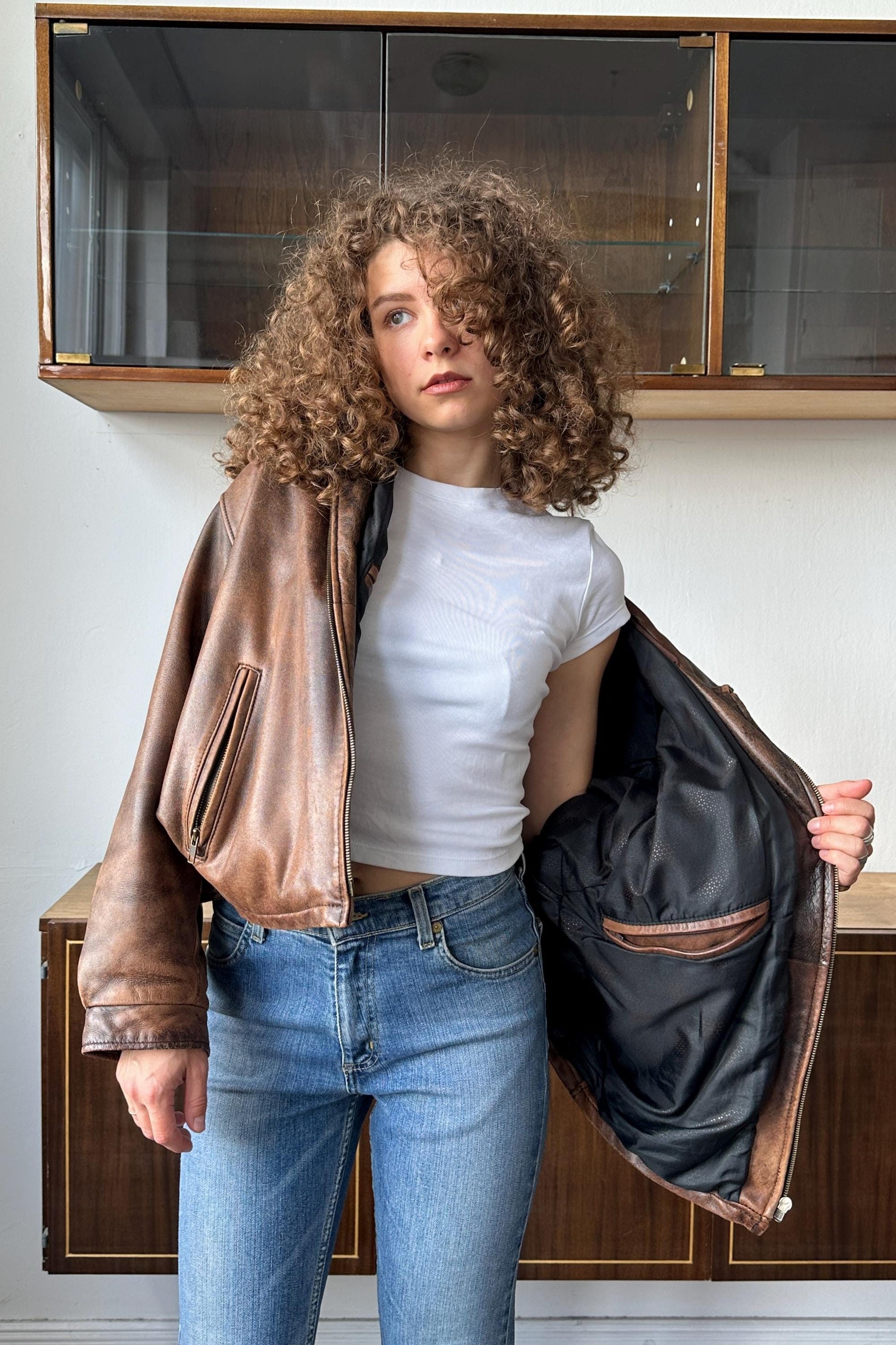 A young woman with curly hair stands in front of a wooden cabinet, wearing a brown leather jacket and a white t-shirt.