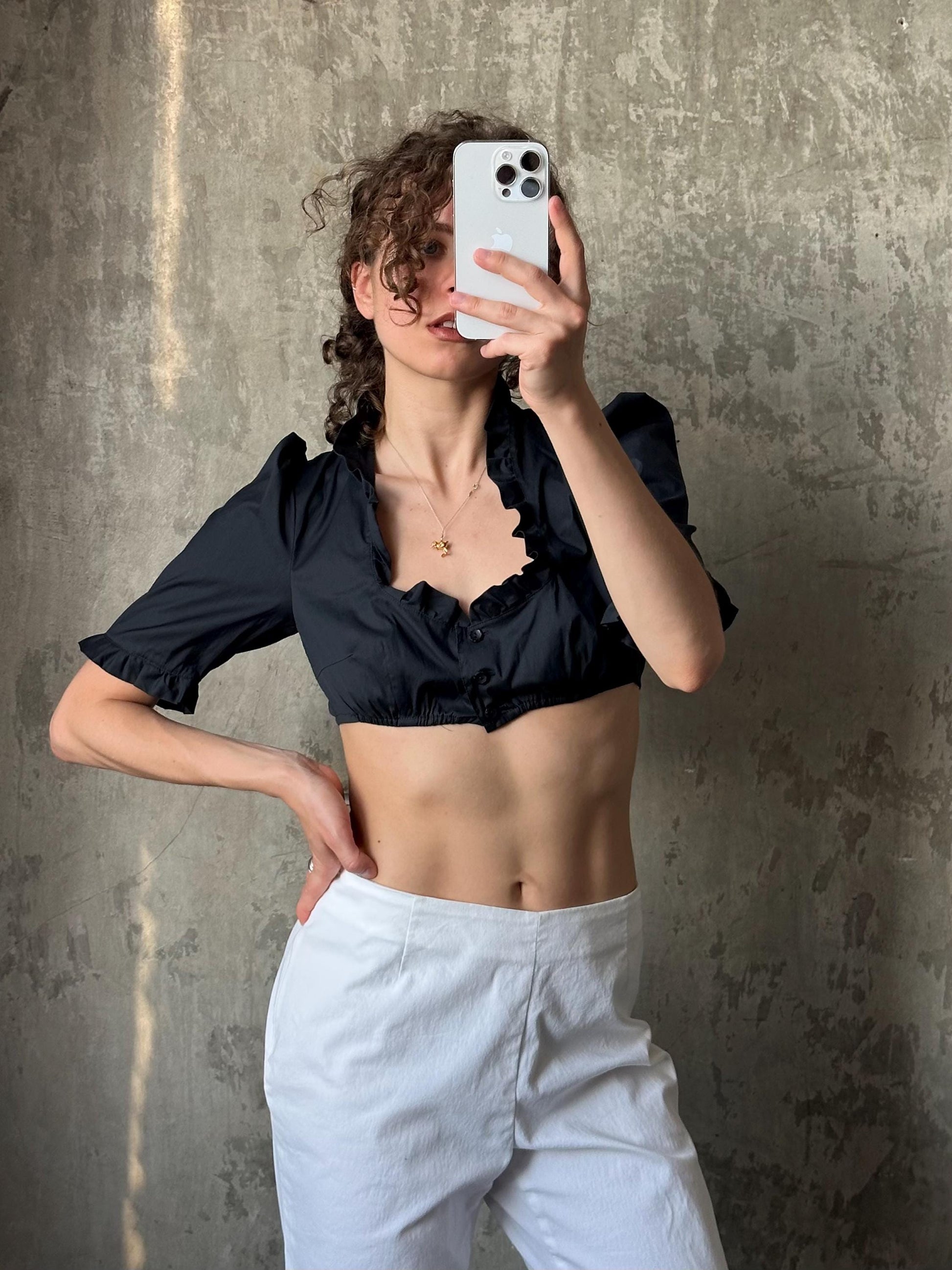 A woman is taking a selfie with her iPhone, wearing a black crop top and white pants, against a textured concrete wall.